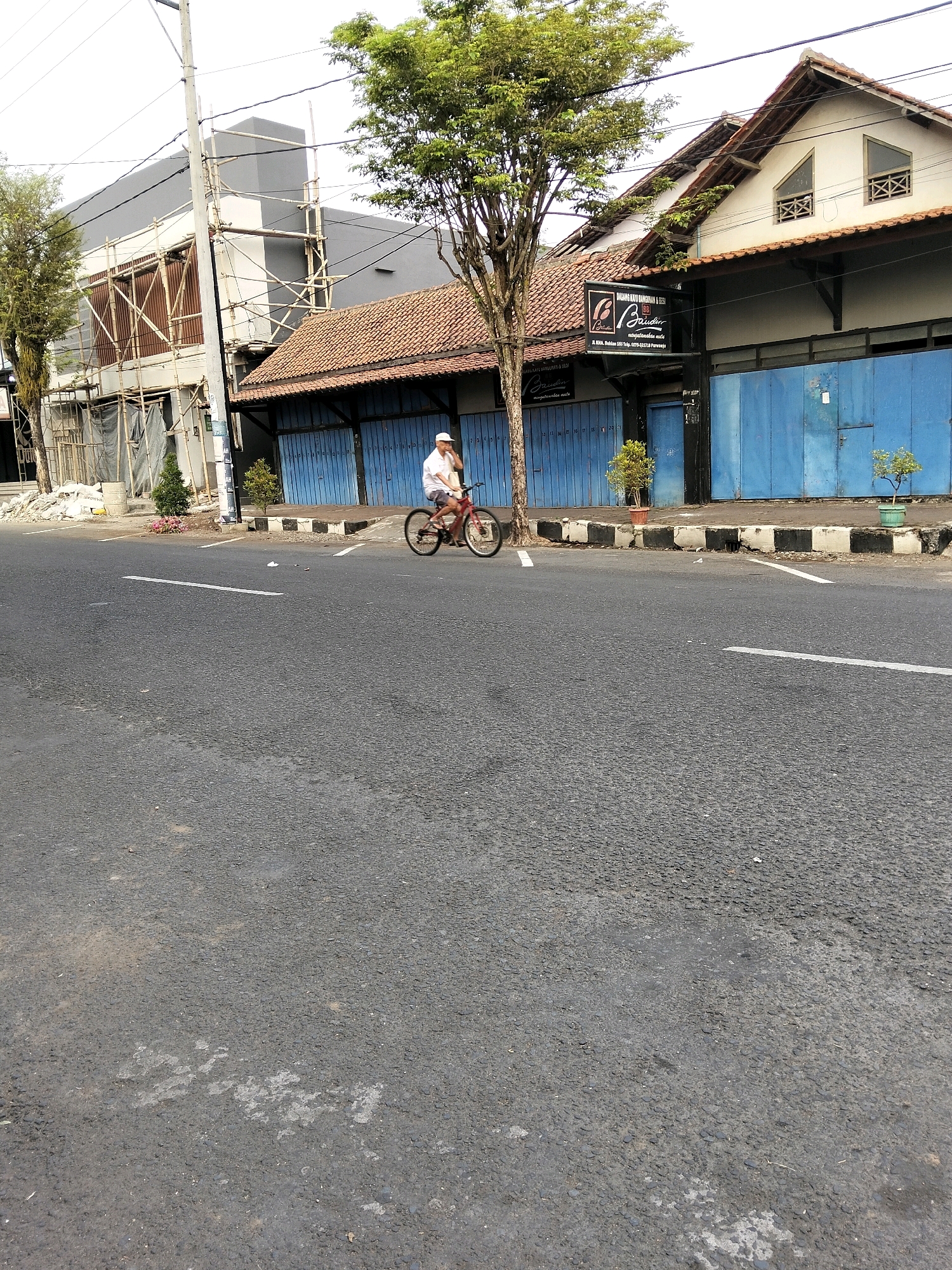 The image shows a street scene in Purworejo, Indonesia. A man is riding a red bicycle on the asphalt road. He is wearing a white t-shirt and a white cap. The road has white lines marking lanes. On the right side of the road, there are several buildings. One building has a brown tiled roof and blue metal doors. Above the doors, there is a sign with text in Indonesian. Another building to the left appears to be under construction, with scaffolding and tarps. A large tree is visible between the buildings and the road. The sky is overcast.