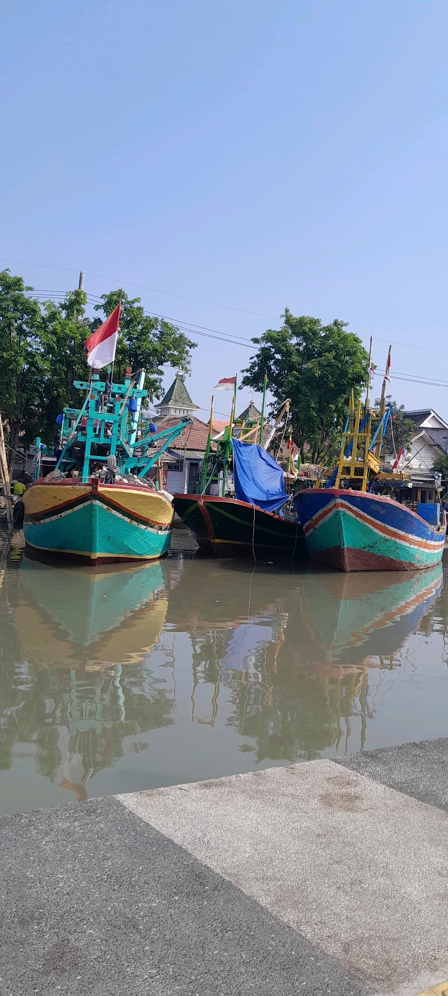 This is a vertical, eye-level, medium shot of several colorful fishing boats docked in a canal in Pasuruan, Indonesia, on a clear day. The boats are lined up, with their reflections visible in the calm, murky water. The boat in the foreground on the left is predominantly teal and yellow, with a red stripe near the top. It features a green structure with white stars. A red and white Indonesian flag flies from its mast. The boat next to it is mostly green with a blue tarp covering part of its deck. The boat on the right is teal and blue with orange and red stripes. It also has a mast with a red and white flag.
Behind the boats, a cluster of buildings with red tile roofs and white walls can be seen, including what appear to be religious structures with distinctive green, pointed roofs. Lush green trees line the bank of the canal, and power lines are visible overhead. The foreground is occupied by a textured concrete walkway, divided by a dark gray section and a lighter gray section, with a shadow cast across it. A small number of people are visible in the background near the boats, appearing to be engaged in activities related to the boats, but their details are unclear. The sky is a clear, pale blue.
