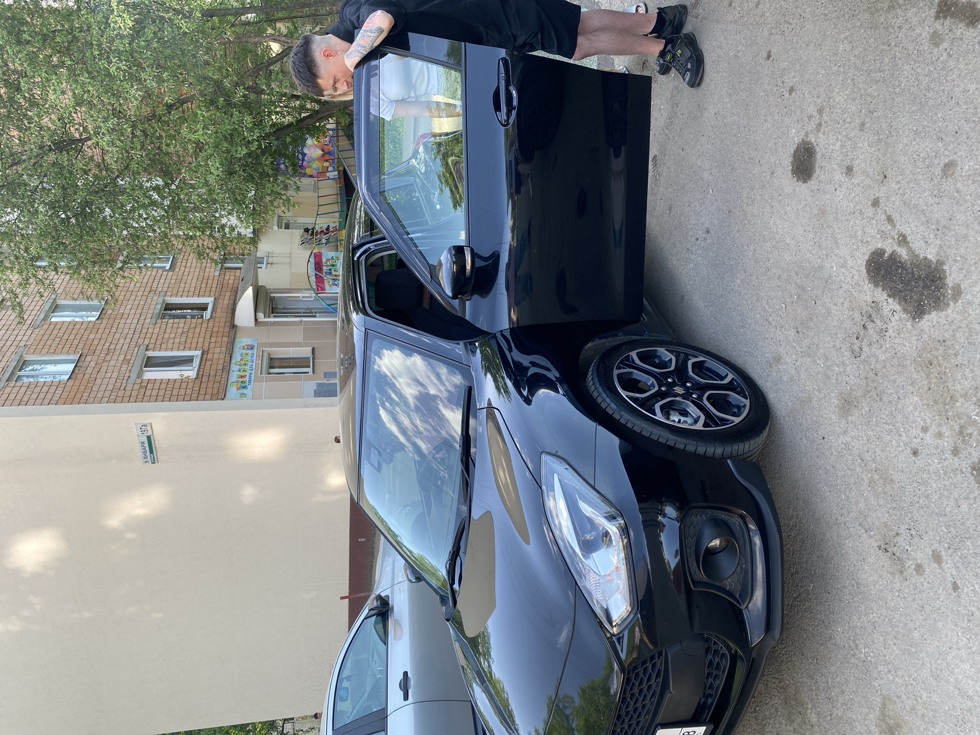 An outdoor, eye-level shot captures a man interacting with a black car on a bright day in what appears to be a residential area in Izhevsk, Russia.
The primary subject is a man with short, styled hair and visible tattoos on his left arm, wearing a black t-shirt, black shorts, and dark athletic shoes. He is leaning casually into the open driver's side door of a shiny black, modern hatchback car, possibly a Suzuki Swift, as indicated by the "SUZUKI" logo on the wheel hub. The car's front bumper shows a partial Russian license plate starting with "88". Another lighter-colored car is partially visible in front of the black car.
In the background, a large, lush green tree stands prominently. Behind the tree and cars are multi-story residential buildings; one is light brown brick, and another is a cream-colored facade. On the cream-colored building, a street sign is clearly visible, reading "9 ЯНВАРЯ 197а" (9 January 197a). Further in the background, a colorful banner or sign with balloons is visible near a building, with Russian text that partially reads "СКАЗКИ ДЛЯ ДЕТЕЙ" (Fairy Tales for Children), suggesting a children's facility nearby.
The ground is concrete pavement, showing some dark stains. The time of day is clearly daylight, indicated by bright, direct lighting and visible reflections of a cloudy sky on the car's windows, suggesting a sunny day. The man's activity appears to be casual interaction with the car, perhaps preparing to enter or exit. The overall scene conveys an ordinary, everyday moment in an urban or suburban setting.