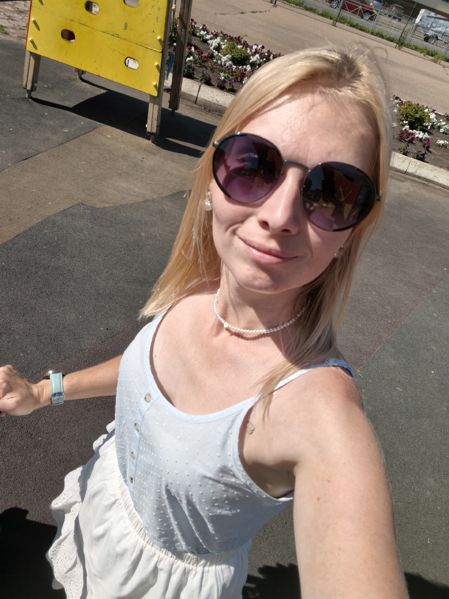 A blonde woman wearing sunglasses and a light blue dress is standing outdoors in what appears to be a playground or park area. The ground is a dark asphalt surface. To her left, a yellow climbing structure with blue accents is partially visible. Behind her, a flower bed with various colored flowers and greenery lines a sidewalk. In the background, there is a street with cars and a fence. The woman is holding something with her left hand, on which she is wearing a light blue watch. She is also wearing a pearl necklace and earrings. The image is taken from a slightly elevated angle looking down at the woman.