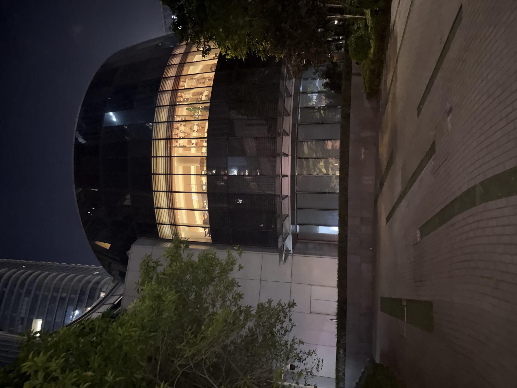 This media file depicts an exterior view of a modern, curved building at night. The building features a glass facade that reveals warm interior lighting, illuminating rows of furniture that suggest an office or commercial space. Adjacent to this structure is another building with a more geometric design, also illuminated from within. The foreground is partially obscured by lush green trees. To the right, a paved pathway with embedded rectangular green spaces leads away from the buildings, creating a sense of an urban plaza or courtyard. The overall impression is one of a contemporary architectural setting in an urban environment. The time of day is clearly night, indicated by the dark sky and artificial lighting. There are no visible people or text in the image. The setting appears to be an outdoor space in an urban area, possibly a business district, given the modern architecture.