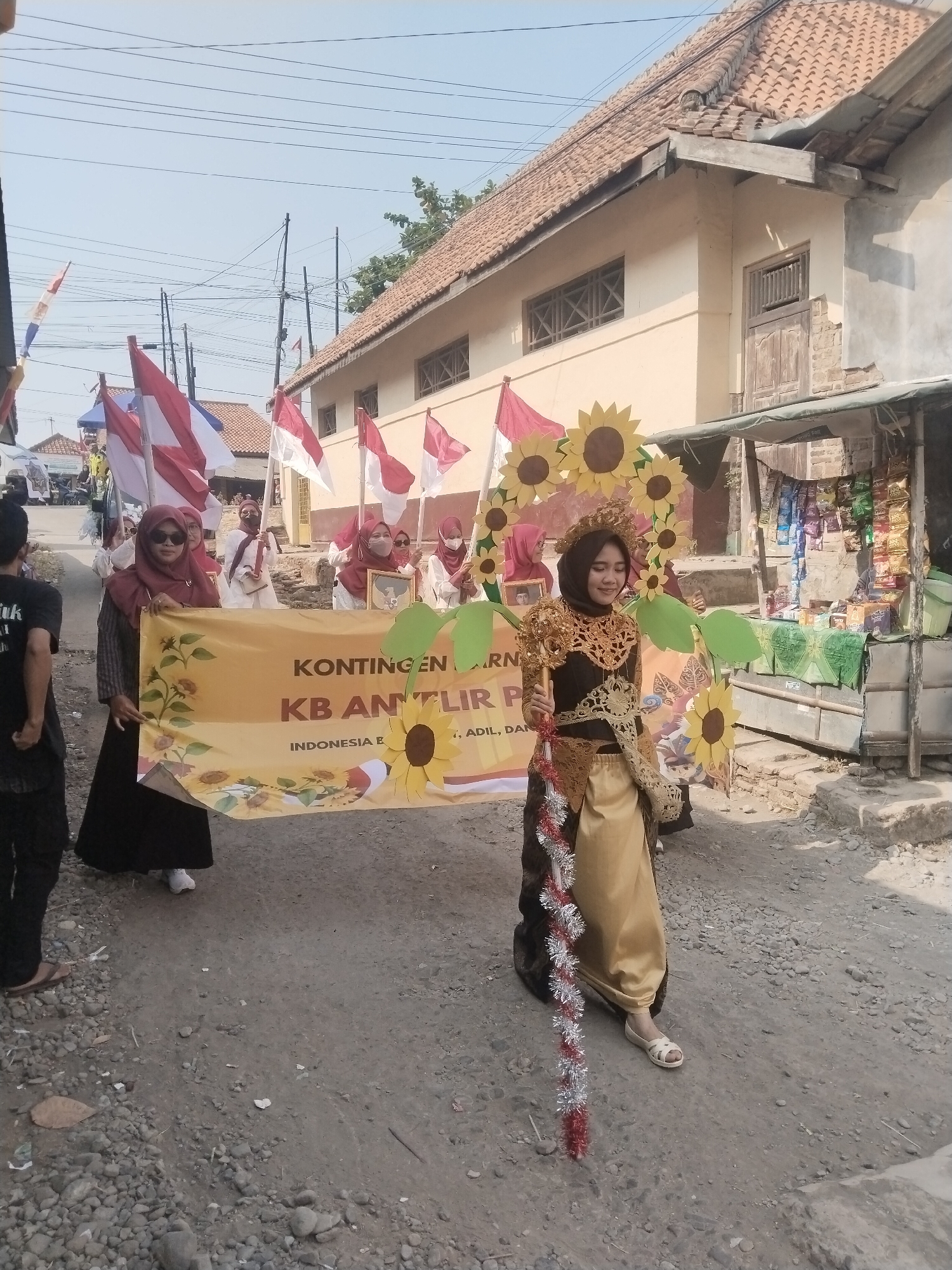 In Jembayat, Indonesia, on a sunny day, a small parade is underway. The street is unpaved, with gravel and debris scattered on the ground. Buildings with terracotta tiled roofs line the street, some showing signs of wear.
At the forefront of the parade, a young woman in a striking costume walks forward. She wears a black and gold ornate top, a golden skirt, and a headdress adorned with what appear to be sunflowers. A string of red and silver tinsel hangs from her costume. She is holding a decorative staff.
Behind her, several women are holding a large yellow banner that reads "KONTINGEN KB ANGLIR PA". The banner is decorated with sunflowers and leaves. The women holding the banner are wearing headscarves, and some are also holding Indonesian flags. Other Indonesian flags are visible throughout the procession. In the background, a small stall is set up selling various packaged goods. The overall atmosphere suggests a festive community event.
