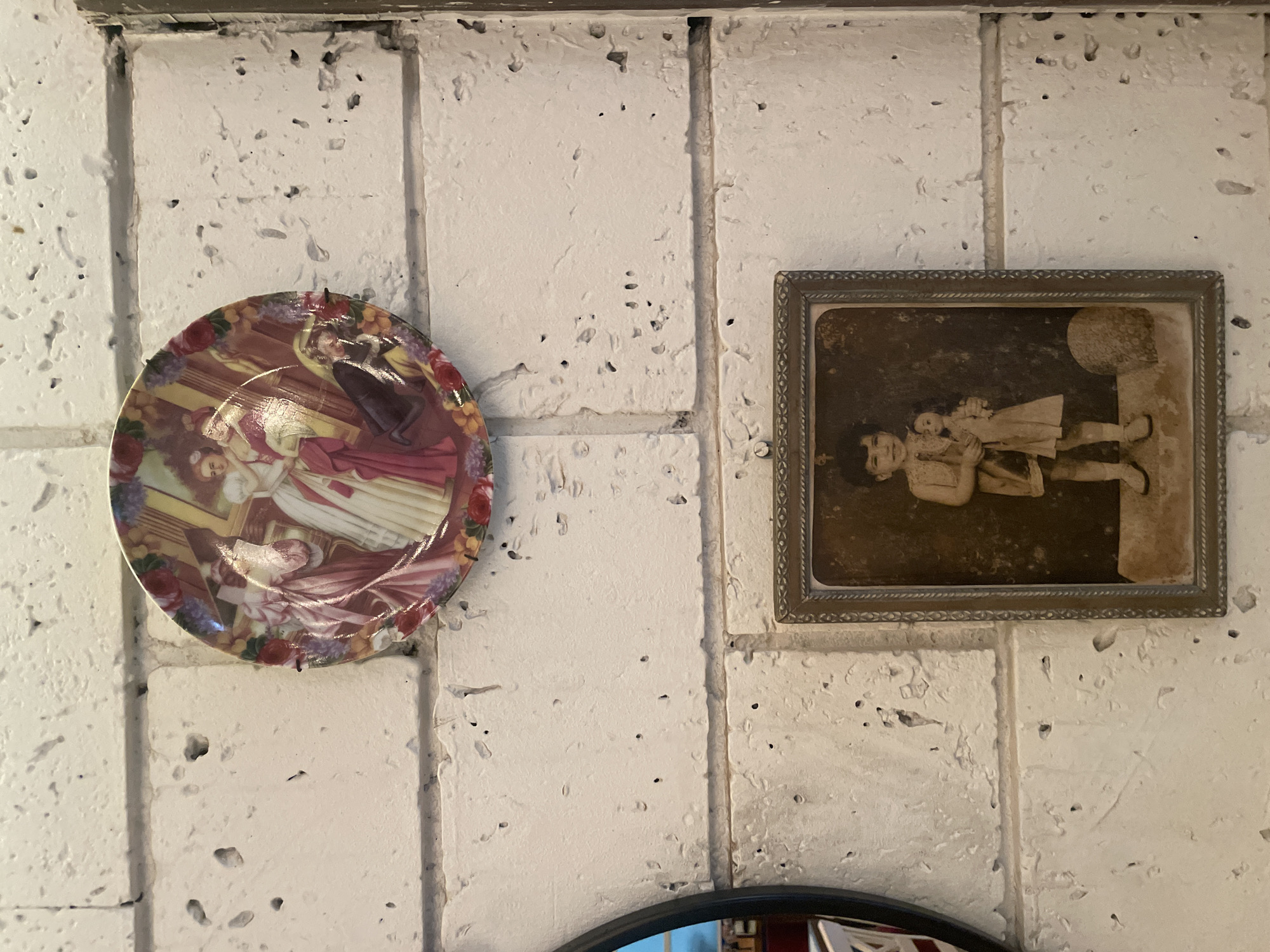 The image shows a decorative plate with a painting of people and a framed sepia-toned photograph of a child holding a doll, both mounted on a white, textured brick wall. The plate is round and colorful, featuring a scene with figures in historical attire. The photograph, framed in ornate metal, depicts a young child with dark, curly hair standing and holding a doll. The wall appears to be exterior or interior, with visible grout lines and a rough, porous surface. A partial view of a round mirror with a black frame is visible on the left side of the image, reflecting part of a room. The overall impression is of a wall decorated with vintage or antique items. The scene does not provide specific clues to determine its location in Mumbai, India.