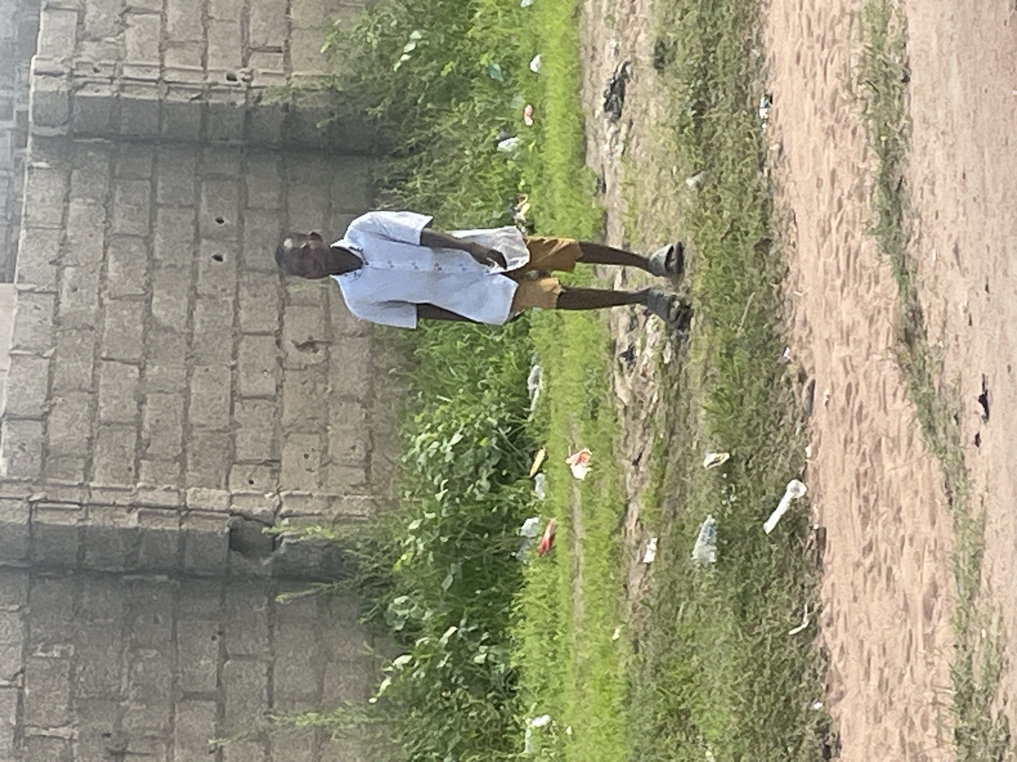 A young boy walks on a dirt path in Jalingo, Nigeria. He is wearing a light blue short-sleeved shirt and tan shorts, with dark sandals on his feet. He is walking from left to right, with his head turned slightly to his left. Behind him is a patch of green grass with scattered litter, and a weathered brick wall. The ground in front of him is a light brown dirt path. The overall scene depicts a simple streetscape with a touch of urban decay.