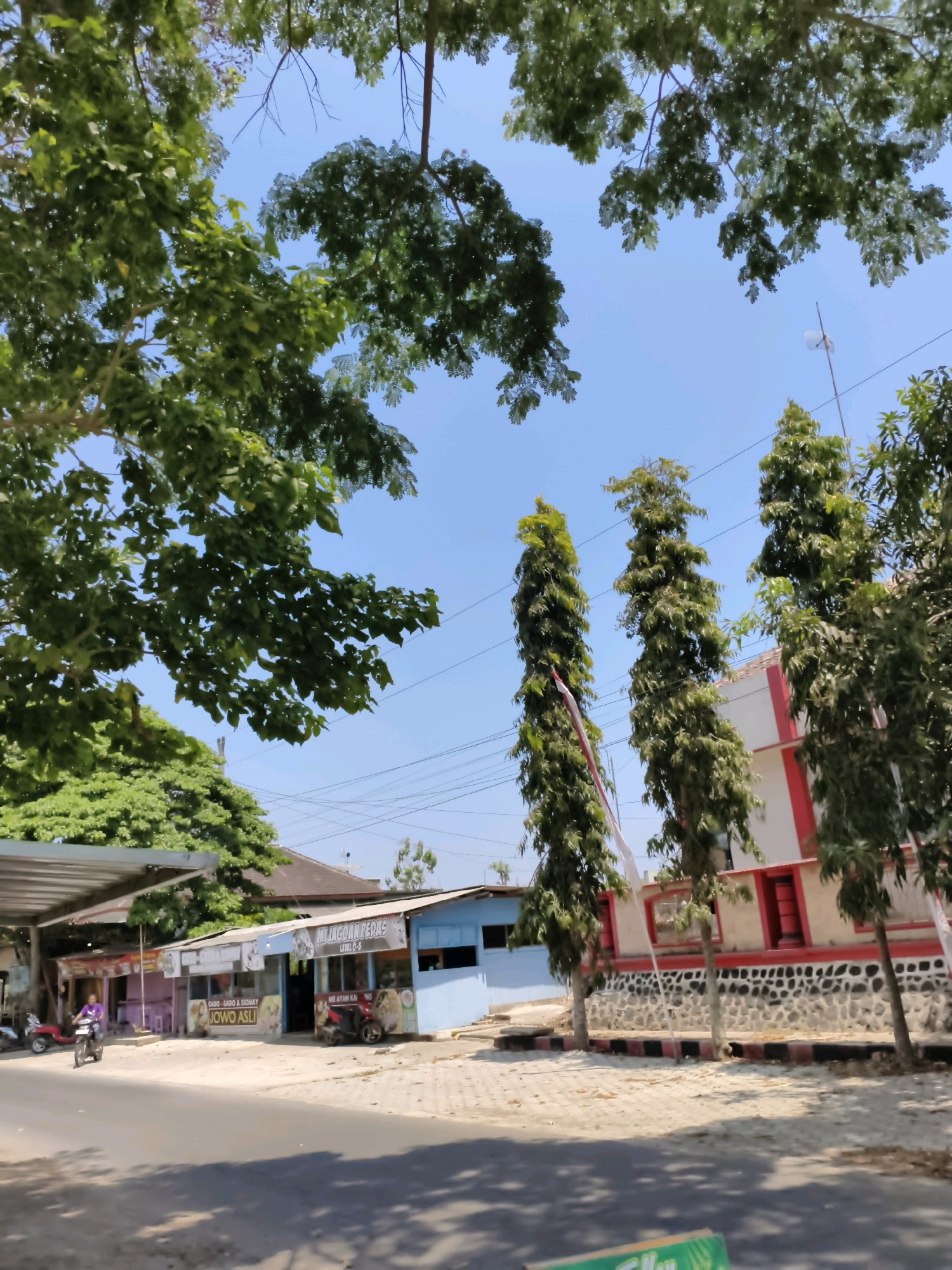 The image depicts a street scene in Puhpelem, Indonesia, under a clear blue sky. Lush green trees with dense foliage frame the top of the image, with their branches reaching towards the sky. In the mid-ground, a row of businesses lines the street, featuring various signs in Indonesian, including "MJ. JAGOAN PEDAS," "GADO-GADO & SIOMAY," and "MI AYAM KAH." A person is seen riding a motorcycle on the left side of the street, in front of a covered outdoor seating area. To the right of the businesses, a building with a white and red facade stands, separated from the businesses by a stone wall. Tall, slender trees are planted in front of this building. The street itself appears to be asphalt, with shadows cast across it from the surrounding trees. Power lines are visible crisscrossing the sky.