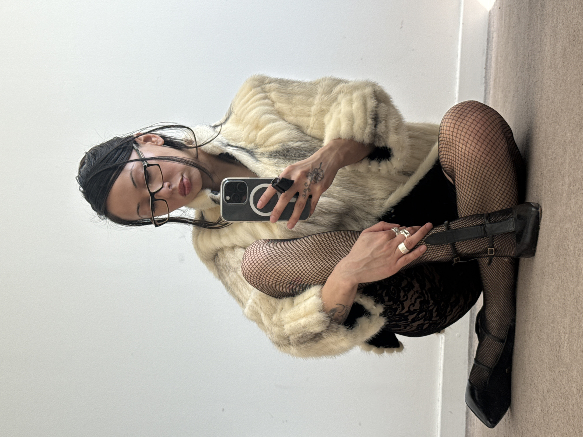 A person is depicted in a close-up, full-body shot, crouched on a light-colored carpeted floor within a brightly lit indoor space. The image is rotated 90 degrees clockwise, orienting the person on their side.
The individual has dark hair, styled away from the face, and wears clear-framed glasses. They are dressed in a plush faux fur jacket, predominantly cream or off-white with distinct black or dark grey markings. Beneath the jacket, they wear black fishnet stockings layered over black lace shorts or a skirt. Black high-heeled shoes with multiple straps and buckles complete the lower ensemble.
The person holds a smartphone, likely an iPhone due to the visible camera array and MagSafe circle, in their left hand, angled towards their face as if taking a selfie or recording. Rings adorn several fingers, including a large silver ring on the right hand and a black band on the left hand. A small, distinctive tattoo resembling a four-leaf clover is visible on the back of the left hand, with another tattoo partially seen on the left forearm.
The setting consists of plain, white or off-white walls, creating a minimalist background. The floor is covered with a light brown or beige carpet. The lighting is even and bright, suggesting an interior environment with good illumination. The individual's expression is subtle, with eyes cast downwards, conveying a pensive or relaxed demeanor. No specific time of day, weather conditions, or visible text (beyond generic phone elements) are discernible in the image.