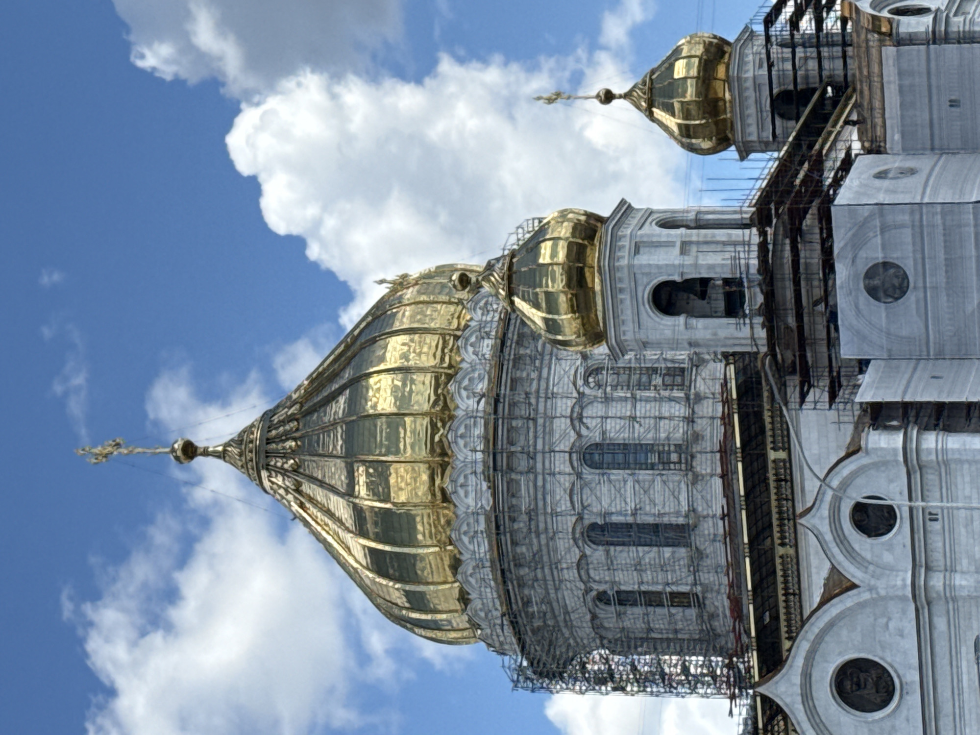 The image shows the domes and upper sections of the Cathedral of Christ the Saviour in Moscow, Russia. The main dome is a large, golden, ribbed structure topped with a cross. To its right, a smaller golden dome sits atop a white, octagonal tower. Further to the right, another golden dome, smaller still, rises from a similar tower. The white stone facade of the cathedral is visible, featuring arched windows and circular medallions. Scaffolding surrounds much of the structure, indicating ongoing construction or renovation. The background is a bright blue sky filled with fluffy white clouds. No people are visible in the image.