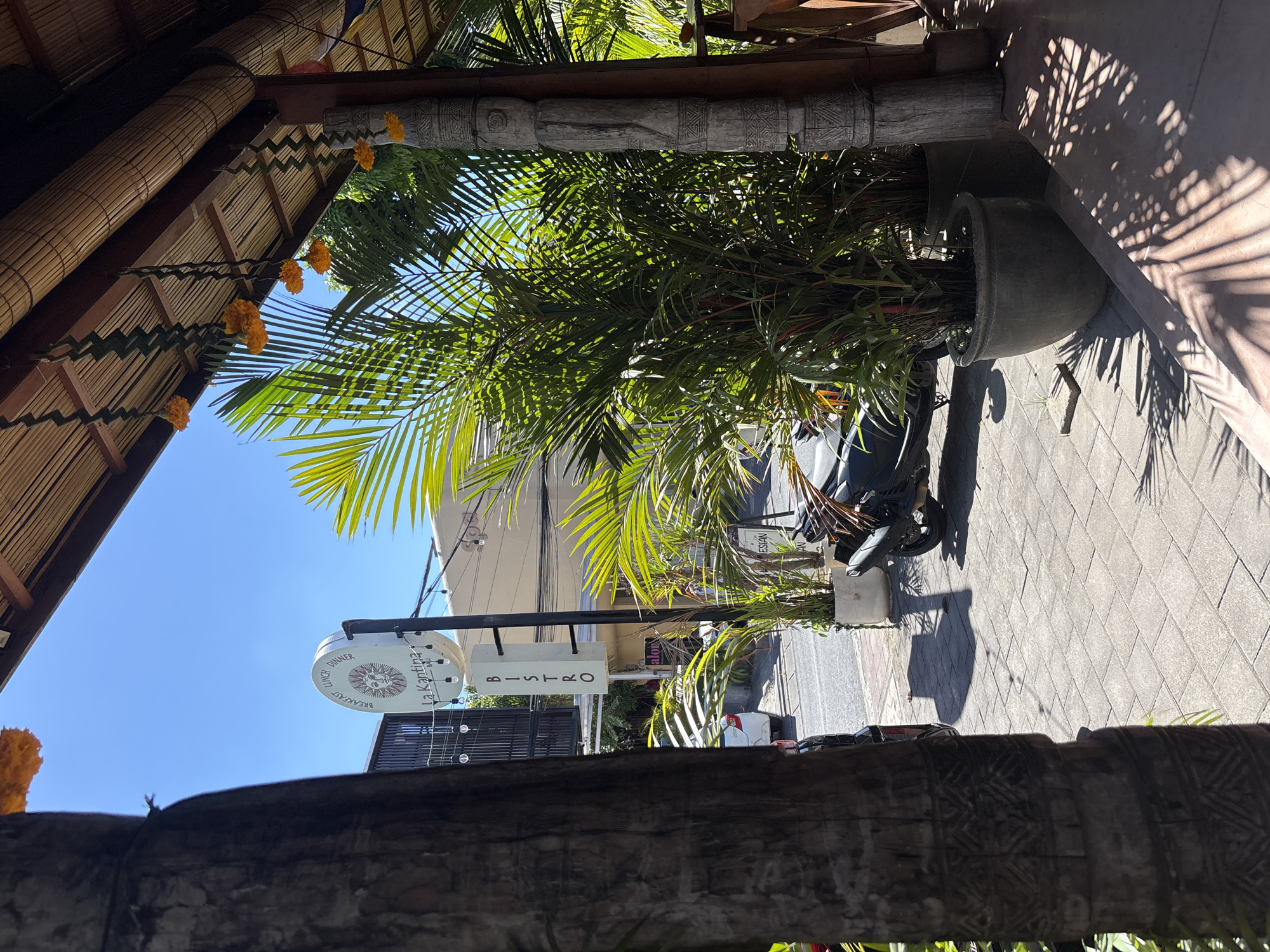 This exterior shot, taken in Pecatu, Indonesia, presents a street scene framed by a bamboo structure adorned with orange flowers. A sign reading "La Kantina BISTRO" is visible. A potted palm is positioned near the street, partially obscuring a parked scooter. The ground is paved with light gray brick. The sky is clear and blue. Light and shadow patterns dapple the ground, cast by the trees and structures overhead.