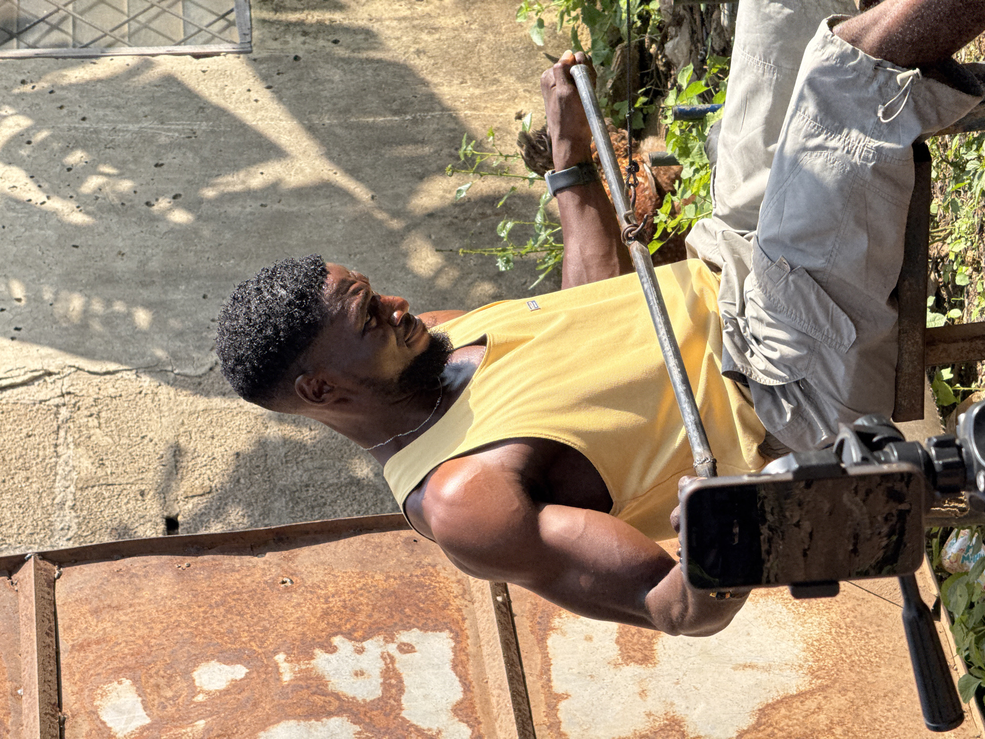 A Black man is sitting outside in Akwanga, Nigeria, engaged in a workout. He is wearing a yellow tank top and light-colored cargo shorts, with a silver necklace and a dark watch on his left wrist. He is holding a metal bar with both hands, his gaze focused downwards on it. A smartphone on a tripod is positioned in front of him, likely for recording his exercise. The background includes a weathered, rusty metal surface on the left and a concrete wall with a window on the right. Green plants and shadows from foliage create a natural, outdoor ambiance.