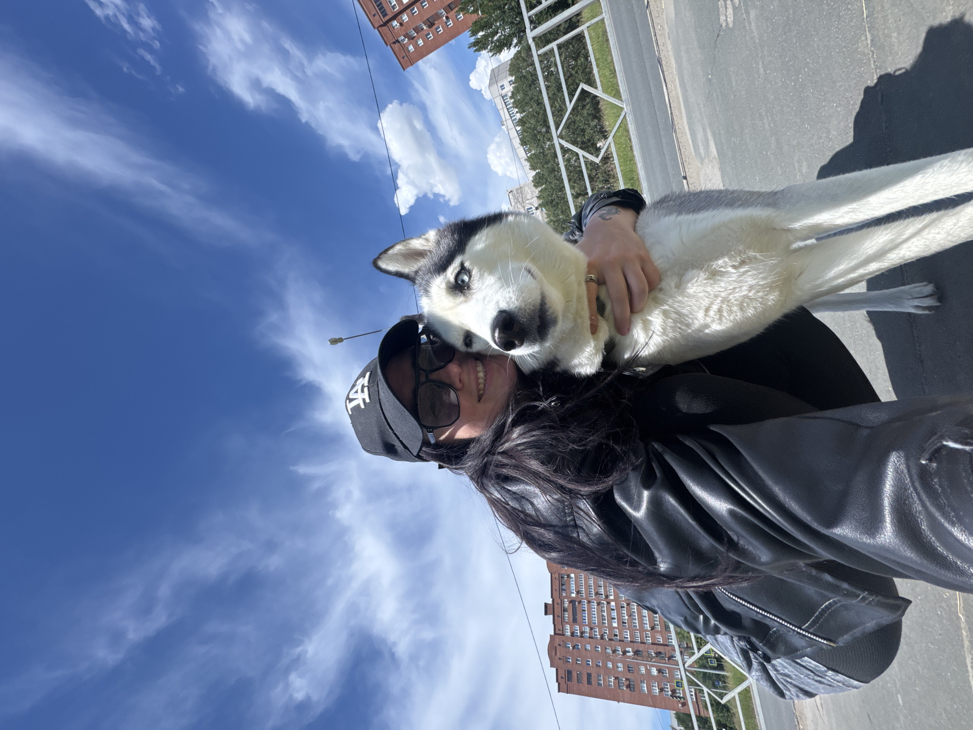 A woman is holding a husky dog in a selfie photo taken outdoors. The woman is wearing a black leather jacket, a black baseball cap with white lettering, and sunglasses. She has dark hair and is smiling. The dog is mostly white with black markings on its head and face. Its eyes are light blue. The background includes a clear blue sky with wispy white clouds, a tall brick building, a metal fence, a street, and a few trees. The location is Unknown, Unknown.