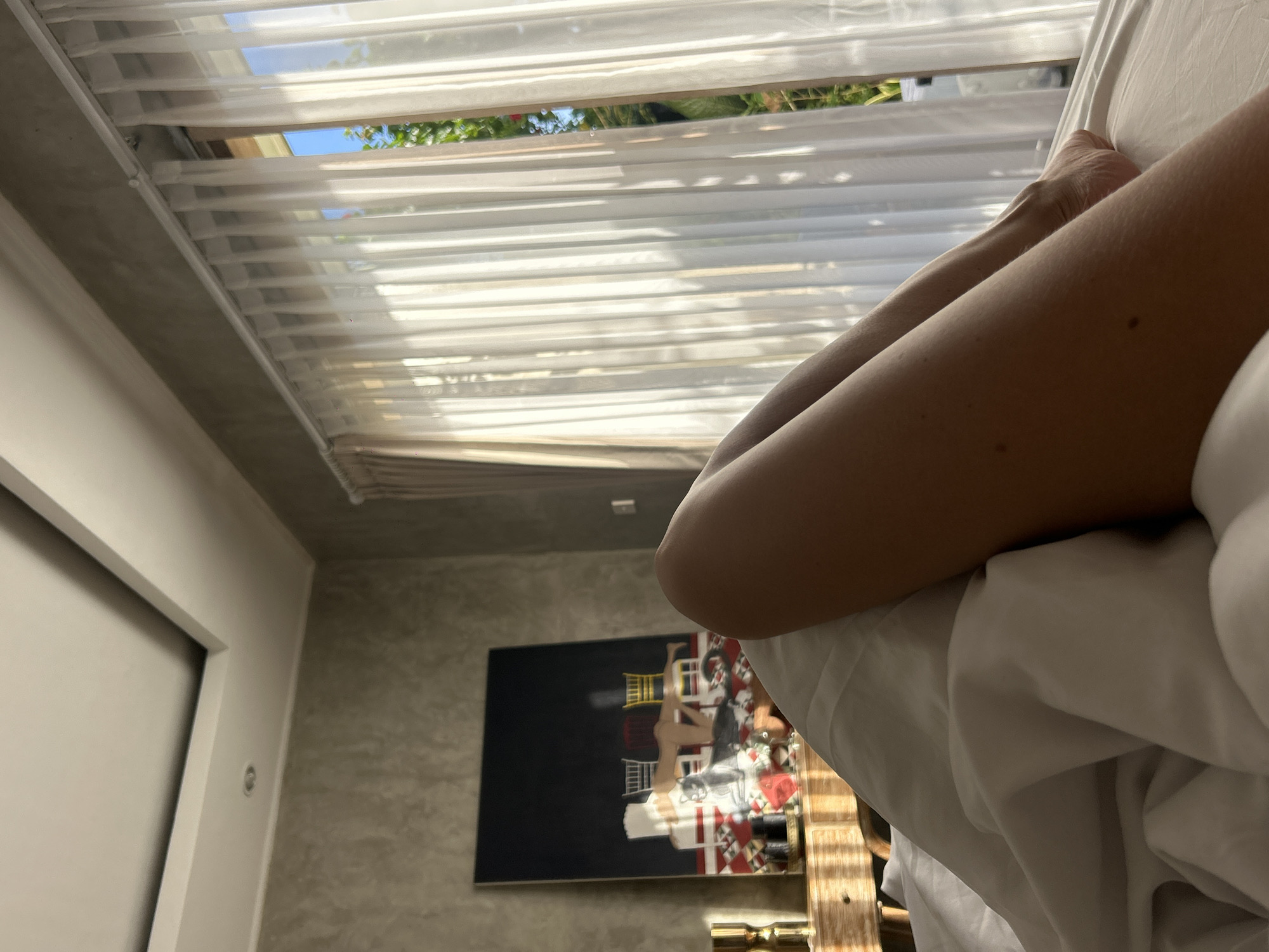 The image shows a person's legs and feet under white bedding, with a view out of a window that is partially covered by white translucent blinds. Outside the window, greenery and a hint of blue sky are visible. In the background, inside the room, there is a painting hanging on a textured gray wall. The room appears to be located in North Kuta, Indonesia.