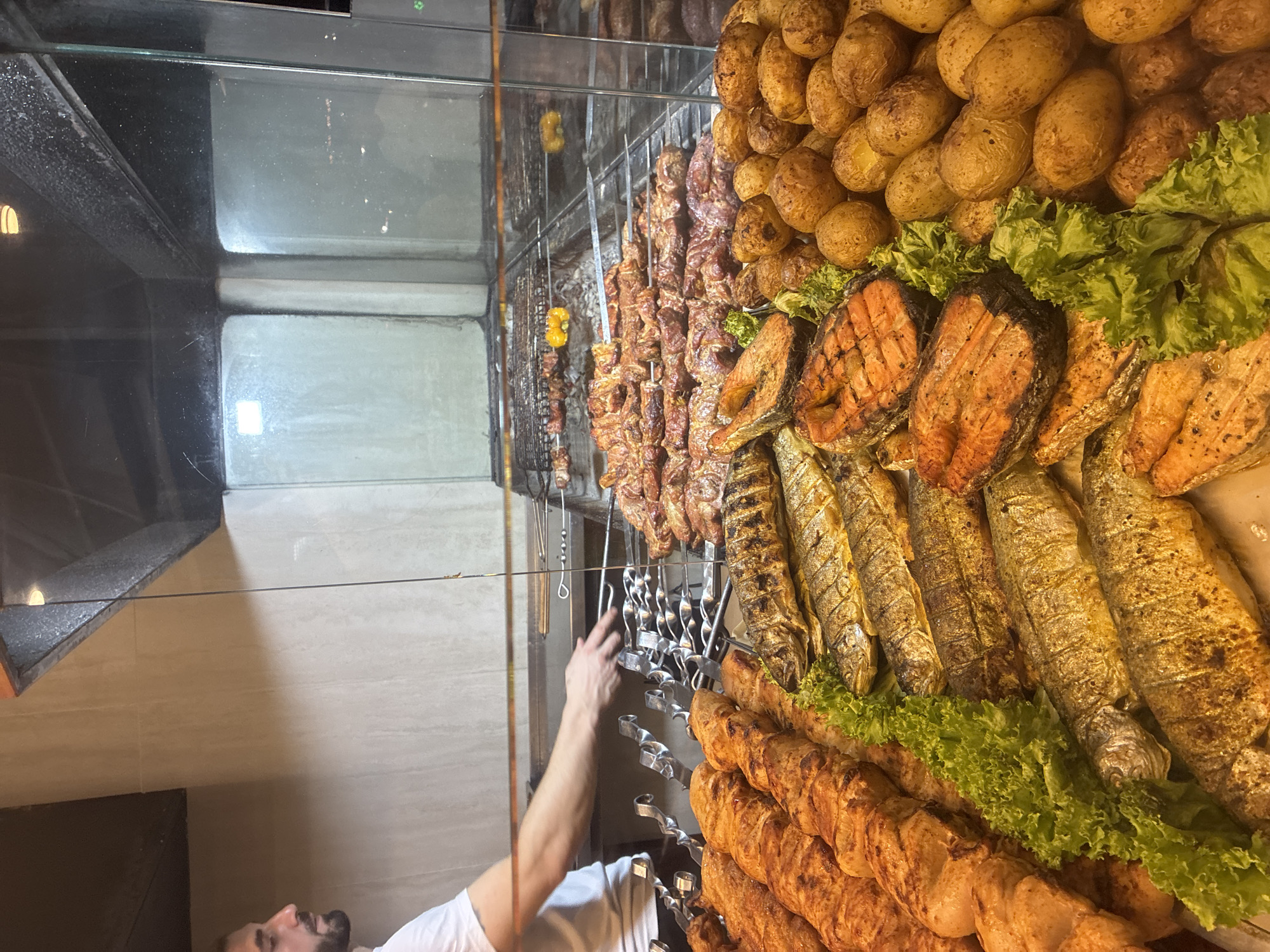 In Sirius, Russia, a man in a white t-shirt is seen from the side, with his right arm extended towards a display of grilled meats and fish. He appears to be a vendor or chef attending to the food. The display case showcases an array of cooked items, including skewers of meat, whole grilled fish, salmon steaks, and small roasted potatoes, garnished with lettuce. In the background, more meat is being grilled over coals in a commercial kitchen setting. The overall environment suggests a restaurant or food establishment specializing in grilled cuisine.
