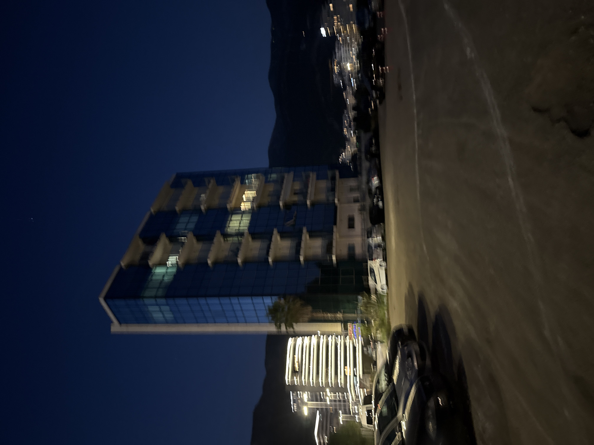 This is a night-time, slightly blurred photograph of a city scene in Boreti, Montenegro. In the foreground, there is a paved parking lot with several cars parked. The cars are dark in color, and their details are indistinct due to the blur and low light. The pavement shows some wear and tear, with cracks and white lines marking parking spaces.
Dominating the mid-ground is a tall, modern building with a facade of blue glass and white balconies. Lights are visible within some of the windows, suggesting the building is occupied. To the left of this building, further in the background, is another multi-story building illuminated with horizontal bands of bright white lights, creating a distinctive illuminated effect.
Behind the buildings, mountains are visible silhouetted against the dark night sky. The sky itself is a deep, clear blue, with a few faint stars visible. The overall impression is of a coastal city at night, with a mix of residential or commercial buildings and natural landscape.