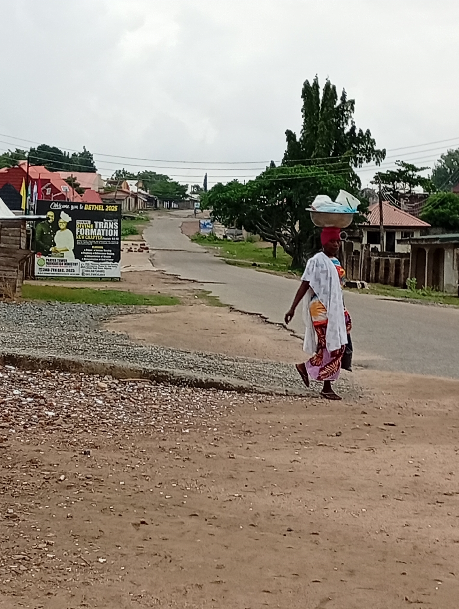 The image depicts a street scene in Lokoja, Nigeria. A woman is walking along the side of the road, carrying a tray of goods on her head. She is wearing a head wrap, a patterned dress, a white cloth draped over her shoulder, and sandals. The road appears to be partially paved, with some sections showing disrepair. Adjacent to the road is a dirt and gravel shoulder. A large sign is visible on the side of the road, advertising a religious event or organization called "Bethel 2025." The background shows a residential area with houses and trees. The sky is overcast.