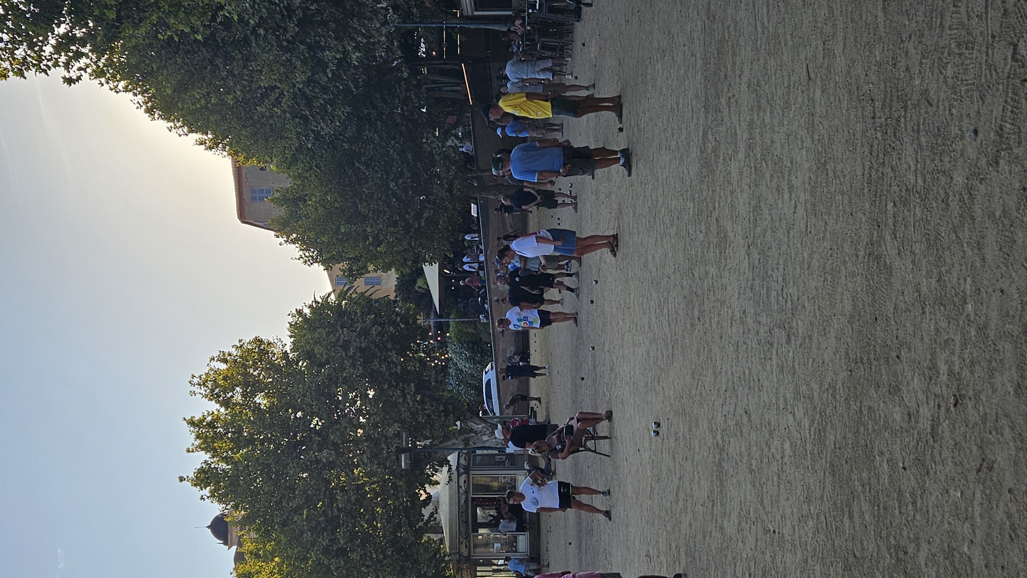 The image depicts a sandy outdoor area with people playing a game of boules, likely a pétanque court, in L'Île-Rousse, France. Several individuals are scattered across the court, some actively engaged in throwing boules, while others observe or stand by. In the background, there are trees, buildings, and a kiosk. The sunlight suggests it is late afternoon or early evening. The ground is sandy and uneven, with several boules visible on the surface. People are dressed casually in t-shirts and shorts.