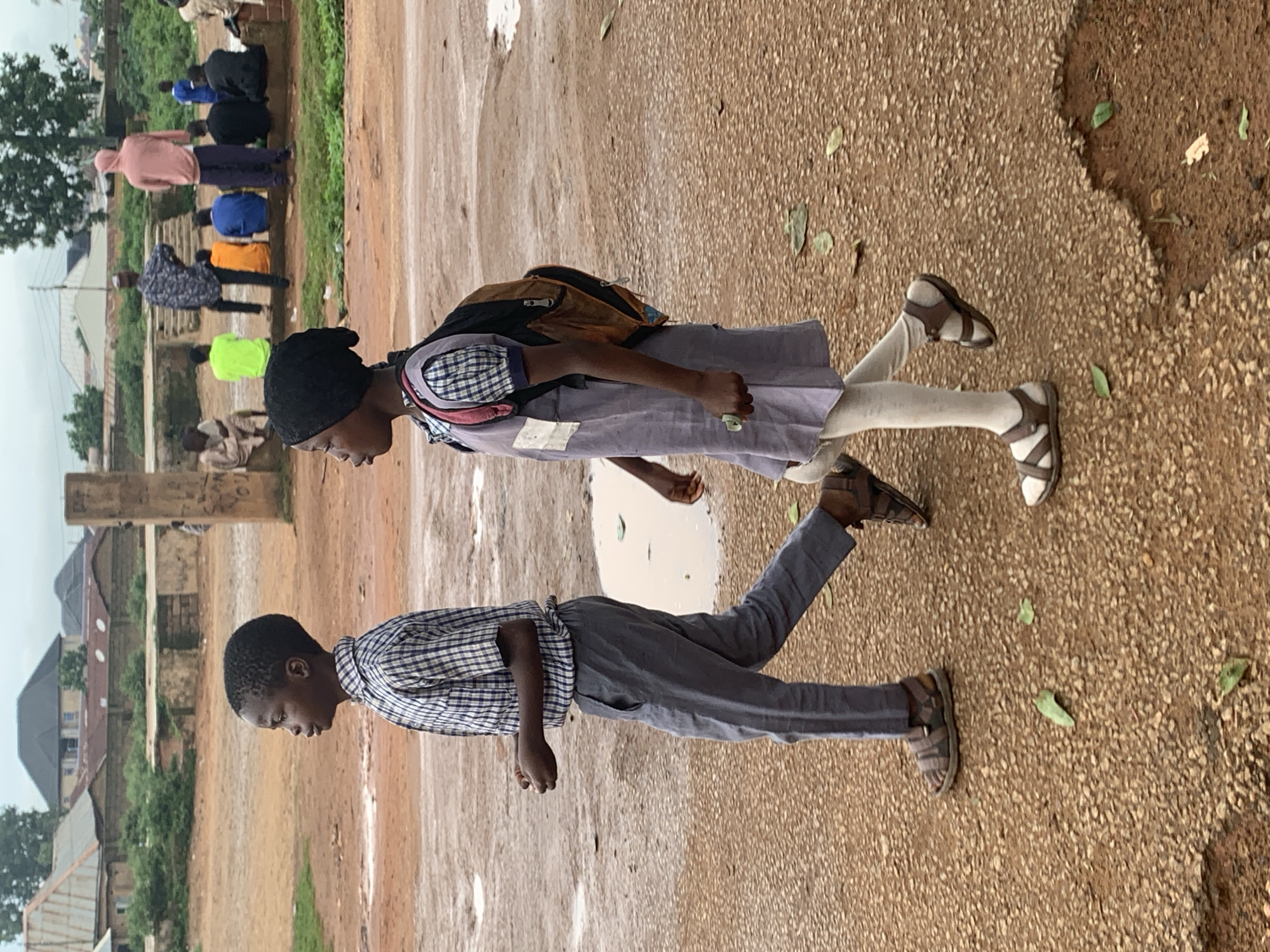 Two children, a boy and a girl, walk on a wet, gravelly ground in Bukuru, Nigeria. The boy, on the left, wears a blue and white plaid shirt and gray pants, with brown sandals. He is looking to the left. The girl, on the right, wears a gray dress over a blue and white plaid shirt, white tights, and brown sandals. She carries a brown backpack and is looking forward. In the background, several other people are seated and standing near a building and a concrete structure, with more buildings and trees visible further in the distance under an overcast sky. The ground is uneven and appears to have puddles, indicating recent rainfall.