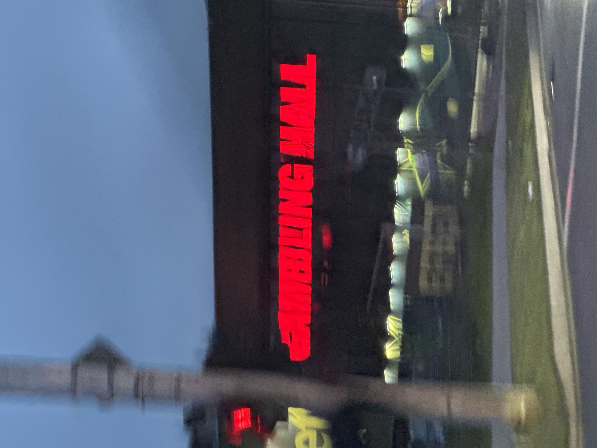 The image depicts a neon sign reading "GAMBLING HALL" in red, affixed to a dark-colored building. The sign is vertically oriented. In the background, blurred lights suggest outdoor structures, possibly construction equipment or lighting. A utility pole is visible in the foreground, obscuring part of the view. The sky is a muted blue, indicating twilight. It is not possible to determine the location in the city of Sofia, Bulgaria from the image.