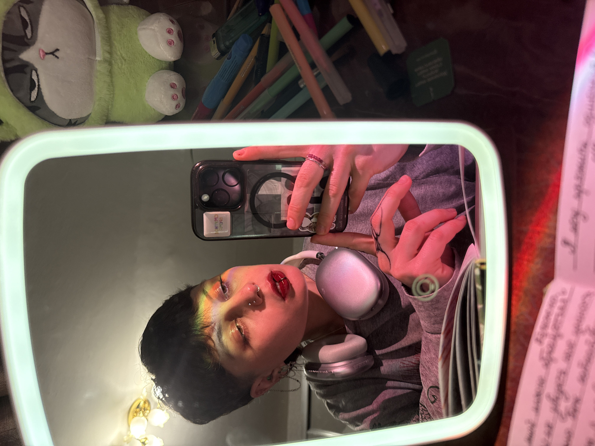 A selfie of a person is reflected in a square mirror with a light. The person is wearing a gray hooded sweatshirt and silver headphones around their neck. The person has piercings on their nose and a decorative make-up around the eye area. They are holding a phone to take the picture in the mirror. On the background of the mirror, there are pens and pencils, a green plush toy. On the right side there is a blurred notebook. Location: Unknown, Saint Petersburg, Russia is not evident in the photograph.