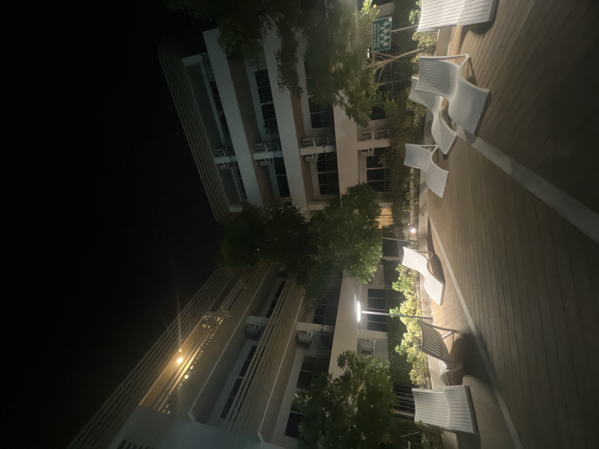 This media file depicts an outdoor seating area at night, adjacent to a multi-story building. The scene features several white, modern chairs arranged on a wooden deck. Trees and foliage are interspersed throughout the area, with some greenery visible behind the chairs and extending towards the building. The building itself appears to be a hotel or dormitory, with visible windows and balconies. Several air conditioning units are mounted on the exterior walls of the building. A streetlamp or exterior light illuminates parts of the deck and building.
The time of day is clearly night, indicated by the dark sky and artificial lighting. There are no people visible in the scene, suggesting a quiet or unoccupied moment. The weather appears to be clear, as there is no indication of rain or fog. The overall atmosphere is serene and somewhat institutional, with the modern design of the furniture and building.
A small green sign with white pictograms is visible in the upper right portion of the image, near the trees. The pictograms appear to represent people in various poses, possibly indicating rules or guidelines for the area. However, the text on the sign is not legible due to the resolution and distance. The image is taken from a slightly elevated perspective, looking down onto the seating area and across towards the building. The perspective is tilted, suggesting the camera was held at an angle.