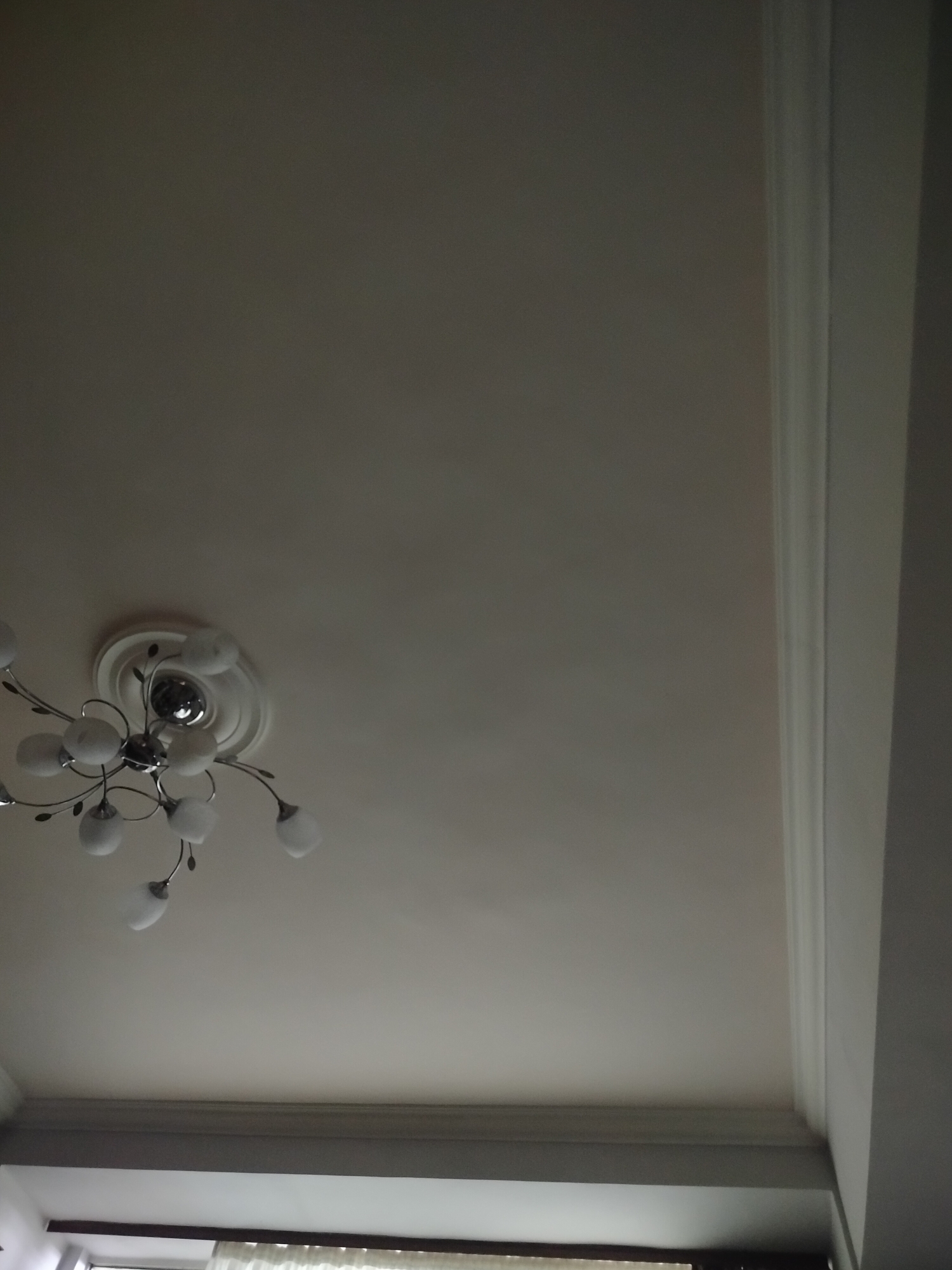 The image shows a view of a ceiling, likely inside a residential building in an unknown city. The ceiling is white with some variation in tone, suggesting lighting differences. A chandelier with multiple light bulbs extends from the ceiling near the left side of the frame. Architectural trim is visible along the right side and bottom edge of the ceiling, meeting at a corner. The bottom portion also reveals a window with what appears to be a curtain. There are no people visible in the image.