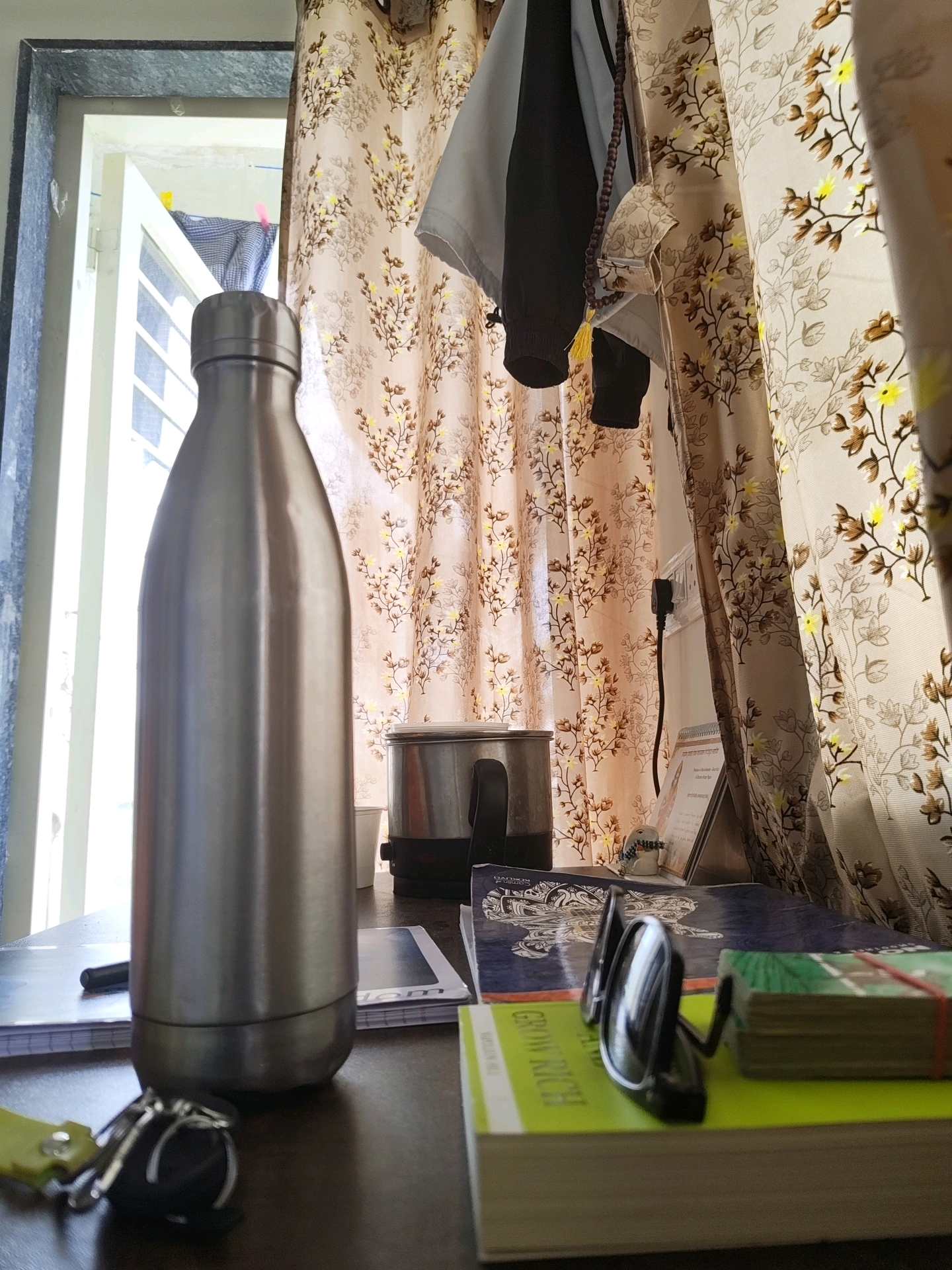 A close-up, low-angle shot captures a collection of objects on a dark wooden surface, possibly a desk, in a room. In the foreground, a silver stainless steel water bottle stands upright, dominating the left side of the frame. To the right of the bottle, a pair of black sunglasses rests on top of a stack of books. The top book is bright green with yellow text, the words "GROW RICH" are partially visible. A stack of smaller books, bound by red elastic bands, sits to the right of the sunglasses. A key ring with a yellow tag and silver keys is visible in the bottom left corner.
In the background, a window with white louvers is partially open, revealing sunlight and an outdoor scene. Floral patterned curtains, in shades of beige and brown with yellow accents, hang to the right, partially obscuring a small appliance, possibly a grinder or blender, with a silver top and black handle. A black electrical cord trails down from an outlet on the wall. A small easel displaying a document is placed between the curtains and the appliance. A prayer bead necklace with a yellow tassel hangs from something above, near the curtains. The overall impression is one of a personal space, likely a home office or study area, in Tathawade, India, as suggested by the mention of the city.
