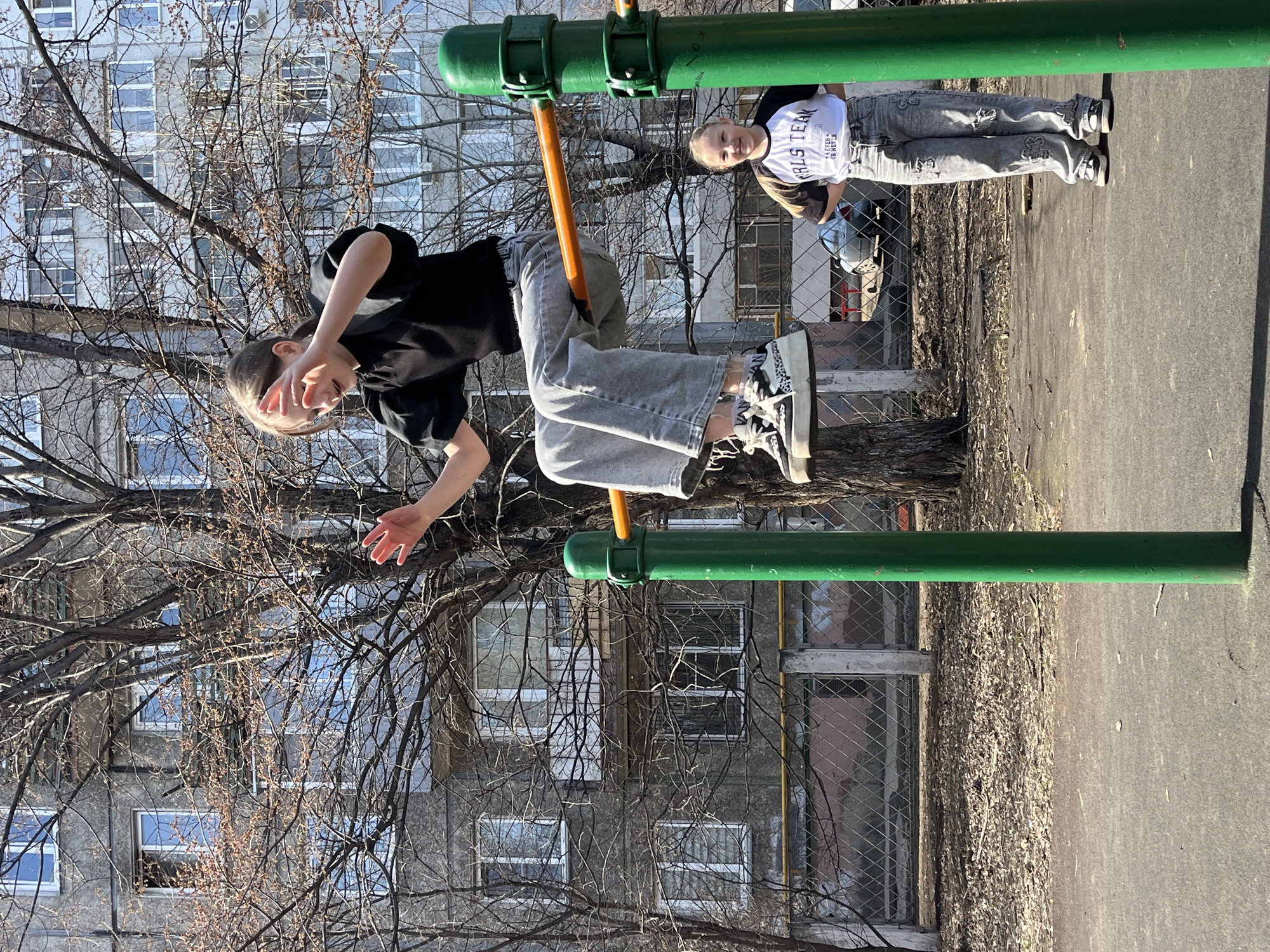 An outdoor, well-lit scene in Krasnoyarsk, Russia, shows two young girls on a sunny day, likely in late winter or early spring due to the barren trees.
The primary subject, a girl with light hair, is playfully interacting with a playground structure on the left side of the frame. She is wearing a black long-sleeved top and light grey, wide-leg jeans adorned with graphic patterns. Her high-top sneakers feature a distinct black and white pattern, possibly animal print. She is hanging from or sitting on an orange horizontal bar, which is part of a green metal playground frame. She looks towards the viewer with a playful smile, partially covering her face with one hand while making a V-sign with her fingers.
To her right, a second girl stands on the asphalt ground, also smiling at the camera. She wears a white t-shirt with black sleeves, featuring the text "GIRLS TEAM" and smaller, partially obscured text below it. Her grey jeans also have similar graphic patterns to the first girl's, and she wears dark sneakers.
The background features a multi-story, grey concrete residential building with numerous windows, typical of older residential architecture. Numerous bare tree branches frame the scene, suggesting the time of year. Strong sunlight casts clear shadows, indicating a bright day. A chain-link fence is visible behind the standing girl. Both girls appear cheerful and engaged with the photographer.