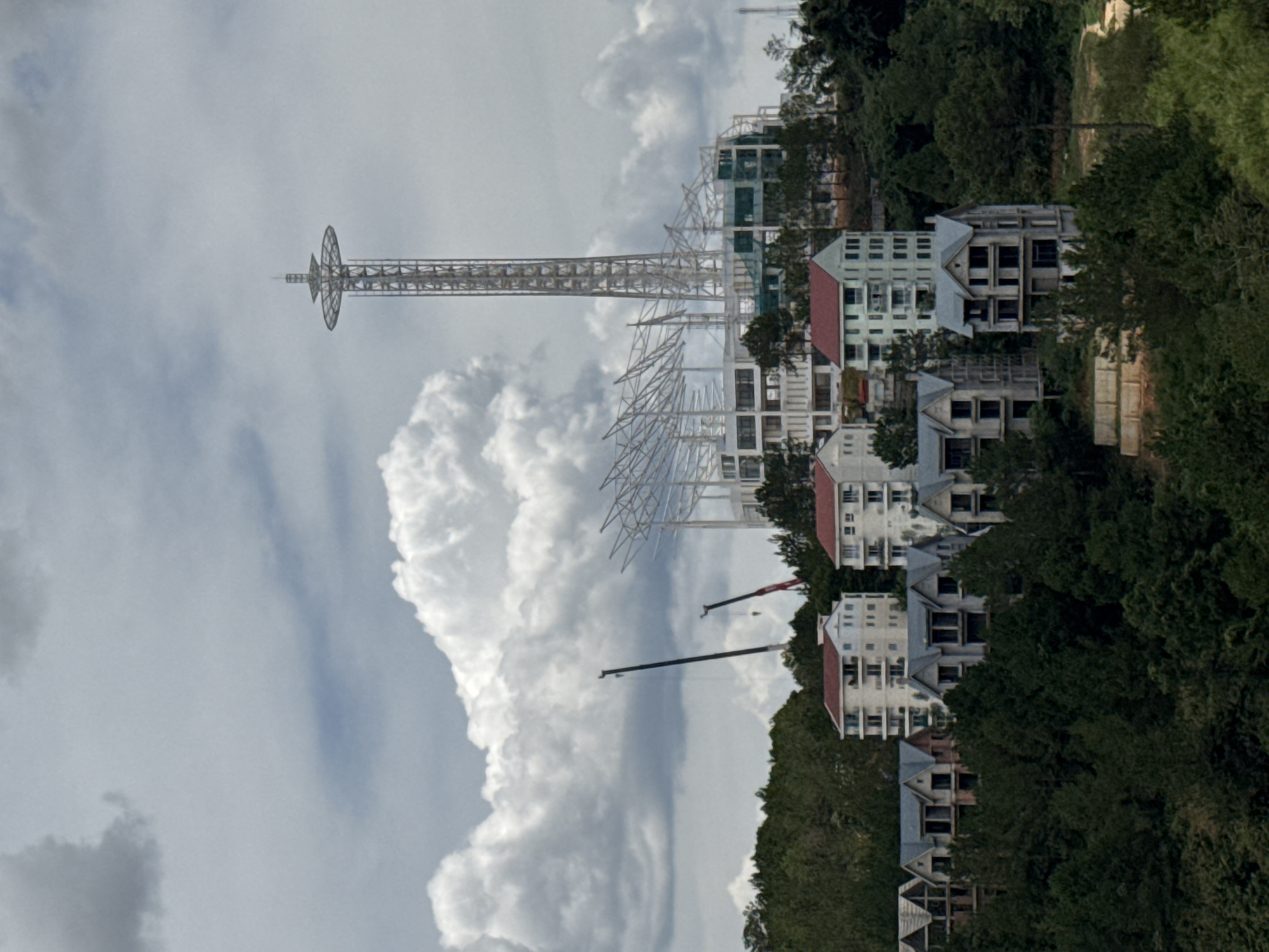 Captured in the city of Da Lat, Vietnam, the scene reveals a partial view of a settlement nestled amongst verdant foliage and partially obscured by cloud formations. A tall, metallic tower pierces the sky, its structural design intricate and distinct. Buildings, some appearing unfinished or under construction, are scattered throughout the scene, their architecture varied and displaying a mix of residential and commercial elements. Cranes are visible, suggesting ongoing construction or development activities. The surrounding environment is lush, with trees and vegetation covering much of the terrain. The sky is dominated by cloudy formations, creating a dynamic and ever-changing backdrop to the scene.