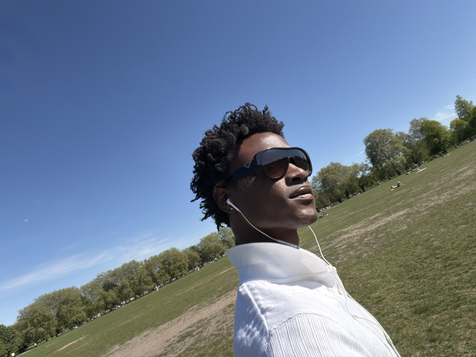 A medium shot shows a person in a white shirt and black sunglasses, with white earphones, in Hyde Park, London, UK. The person's face is tanned and turned upwards. The background shows a large green grassy field with people relaxing, and a line of trees. The sky is blue with some wispy clouds.