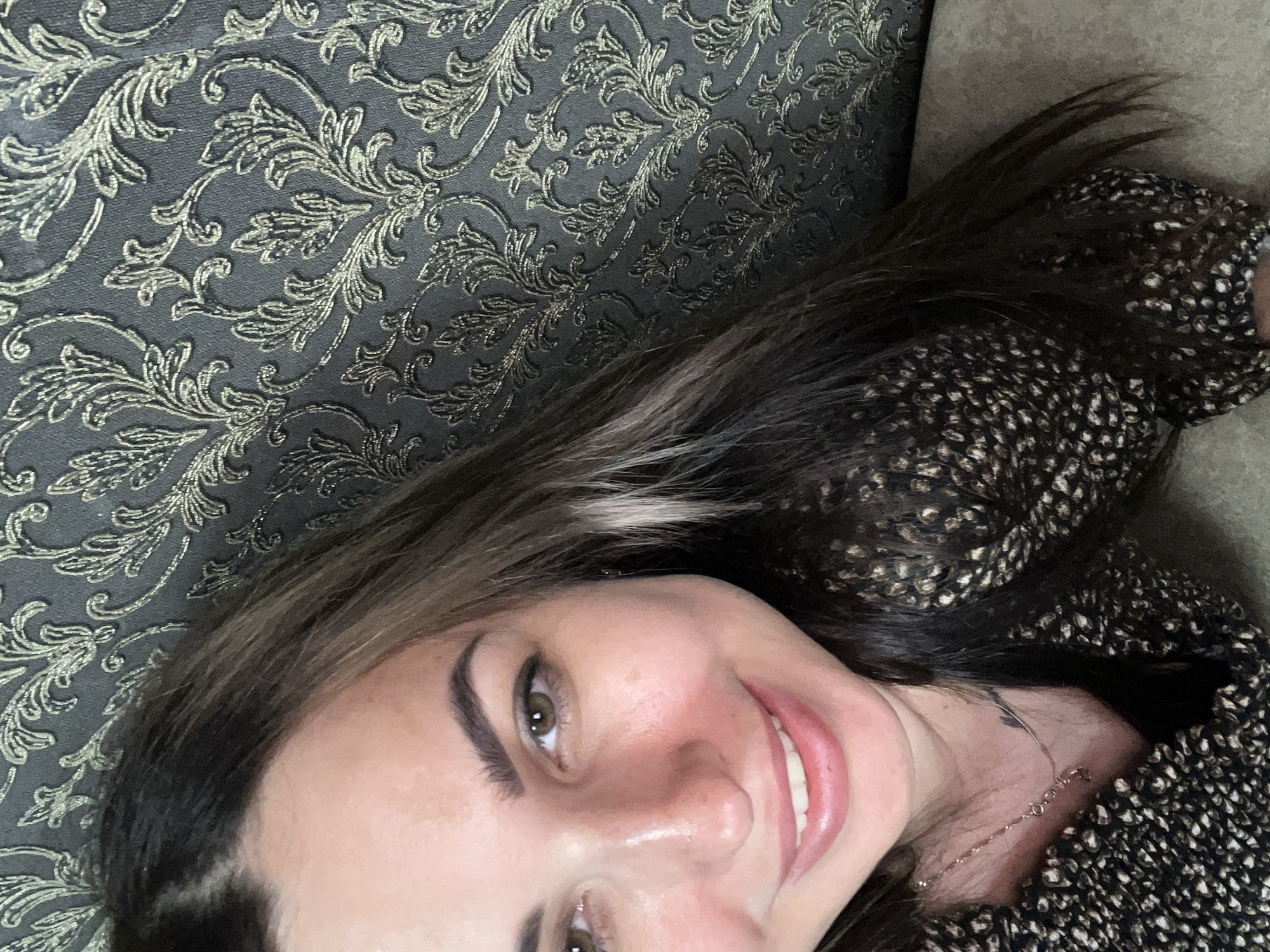 A close-up, head-and-shoulders selfie of a smiling woman, oriented sideways. She has long, dark hair with some lighter streaks, which falls around her face and shoulders. Her eyes are green, and she is wearing a dark, patterned top. The background consists of two different textures: on the left, a dark fabric with a raised, ornate floral pattern in a lighter metallic color; on the right, a plain, textured surface, possibly a couch or cushion, in a neutral tone. The lighting suggests an indoor setting, likely during the day, with no specific time or weather cues. The woman's expression is happy and relaxed. A delicate necklace is visible around her neck, with a small charm or pendant. A faint tattoo is visible on her neck, partially obscured by her hair. The overall impression is casual and personal.
