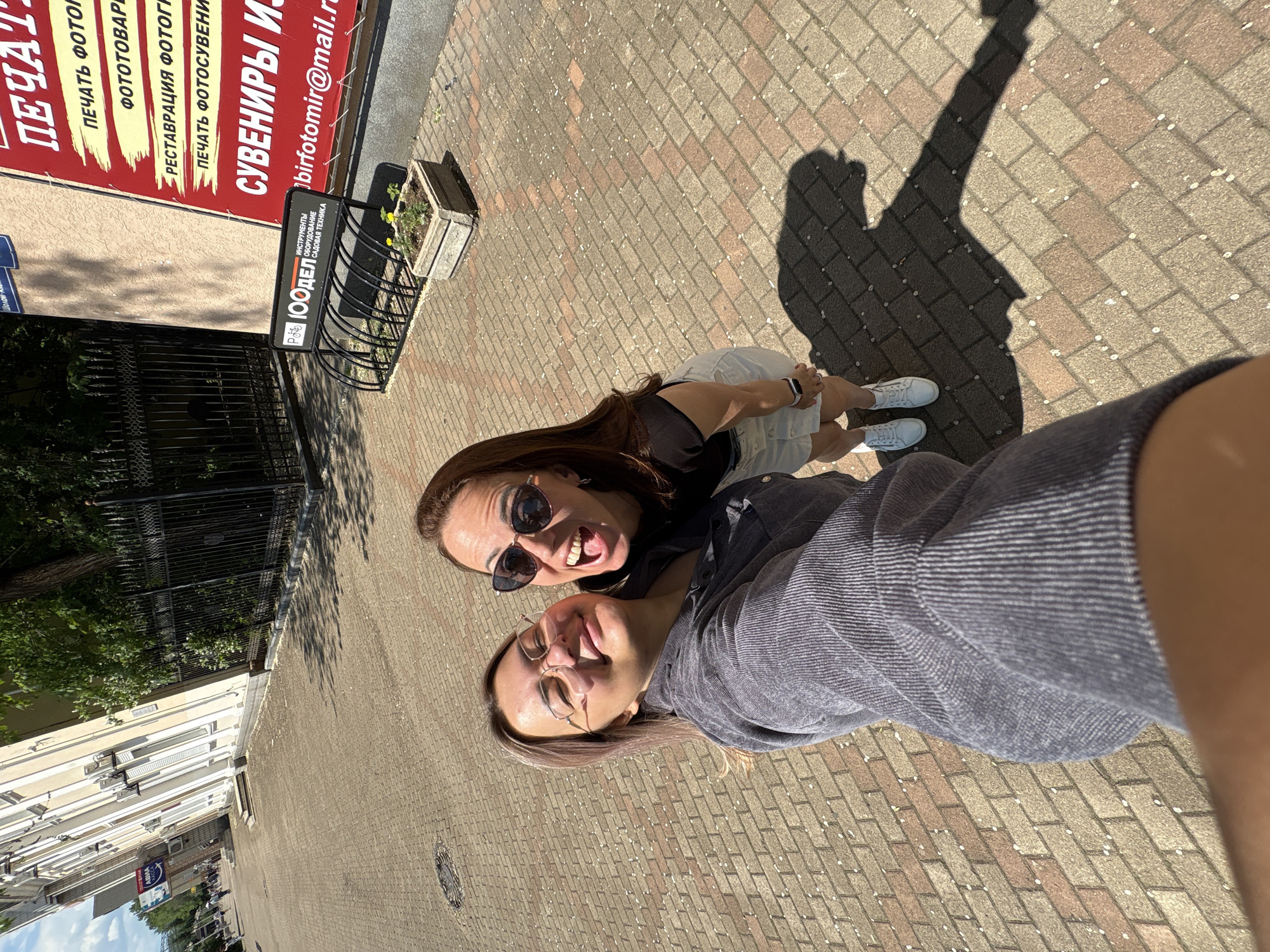 Two women are posing for a selfie on a paved city sidewalk on a sunny day. The woman in the foreground is wearing glasses and a grey corduroy jacket. The woman in the background is wearing sunglasses and a black t-shirt with a white denim skirt. Behind them, a building with white and beige facades and several air conditioning units is visible, along with a black metal fence and a bicycle rack. A red sign with Russian text is attached to the building. The sidewalk is paved with reddish-brown and grey bricks, with scattered white petals. The scene is likely in Birobidzhan, Russia, given the presence of the sign and Cyrillic script.