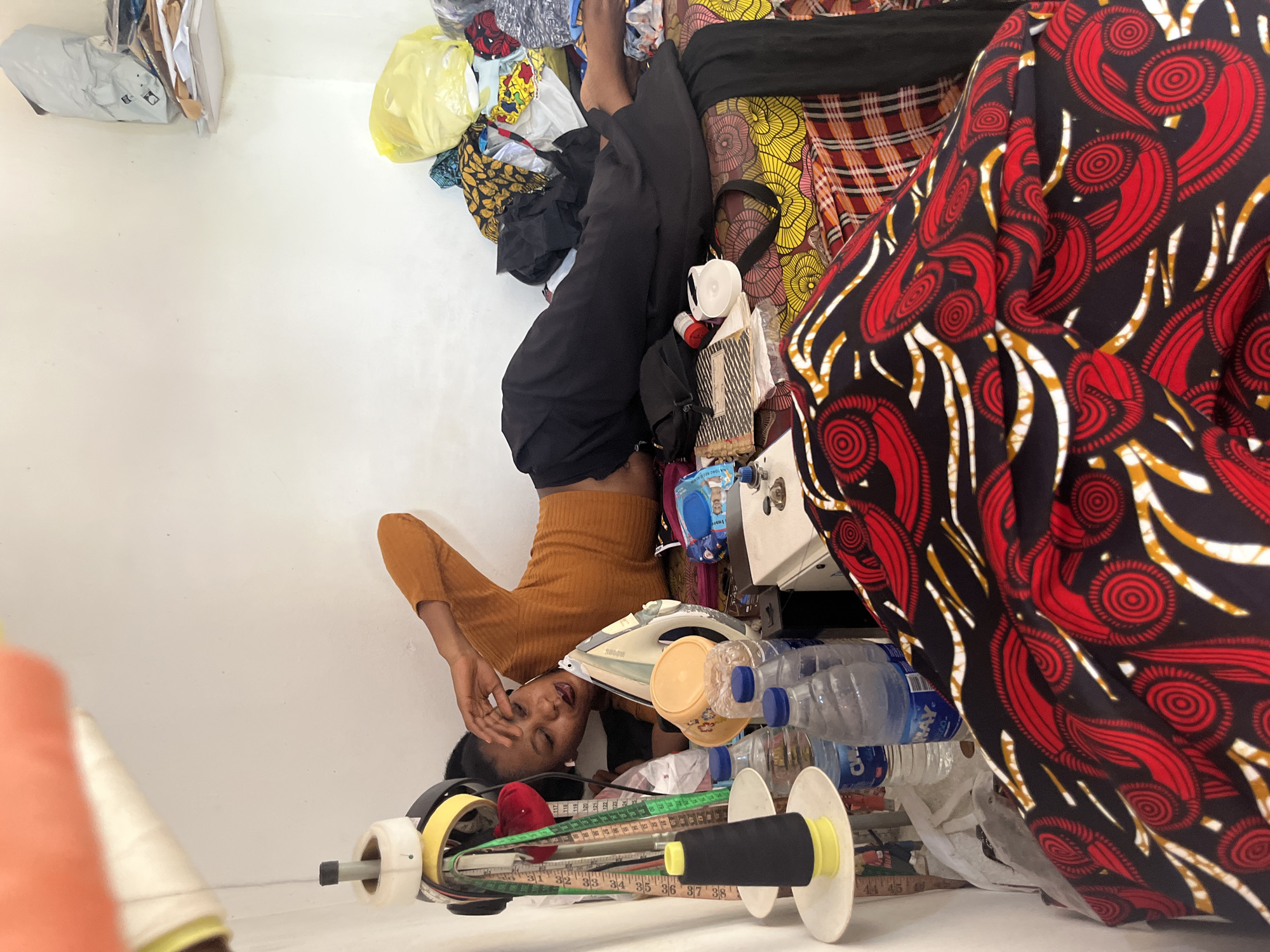 The image depicts a young Black woman in a dimly lit room, likely a tailoring workshop, in Abuja, Nigeria. She is lying on her back with her legs propped up on a pile of colorful African fabrics. Her body is positioned in an unusual and somewhat contorted manner. She is wearing a fitted, rust-colored long-sleeved top and black pants. Her eyes are closed, and her hand is resting on her forehead, suggesting fatigue or distress.
The scene is cluttered with various items related to tailoring. In the foreground, a sewing machine bobbin with black thread is visible, along with measuring tapes. Behind the woman, an iron, plastic bottles of water, and fabric scraps are scattered. The background is filled with more fabric rolls and bags of materials. The overall impression is one of a busy workspace, possibly with a deadline or an overwhelming amount of work. The lighting is artificial, and there are no indications of the time of day or weather. The woman's posture and expression evoke a sense of exhaustion, but also resilience in the face of hard work. There is no visible text in the image.