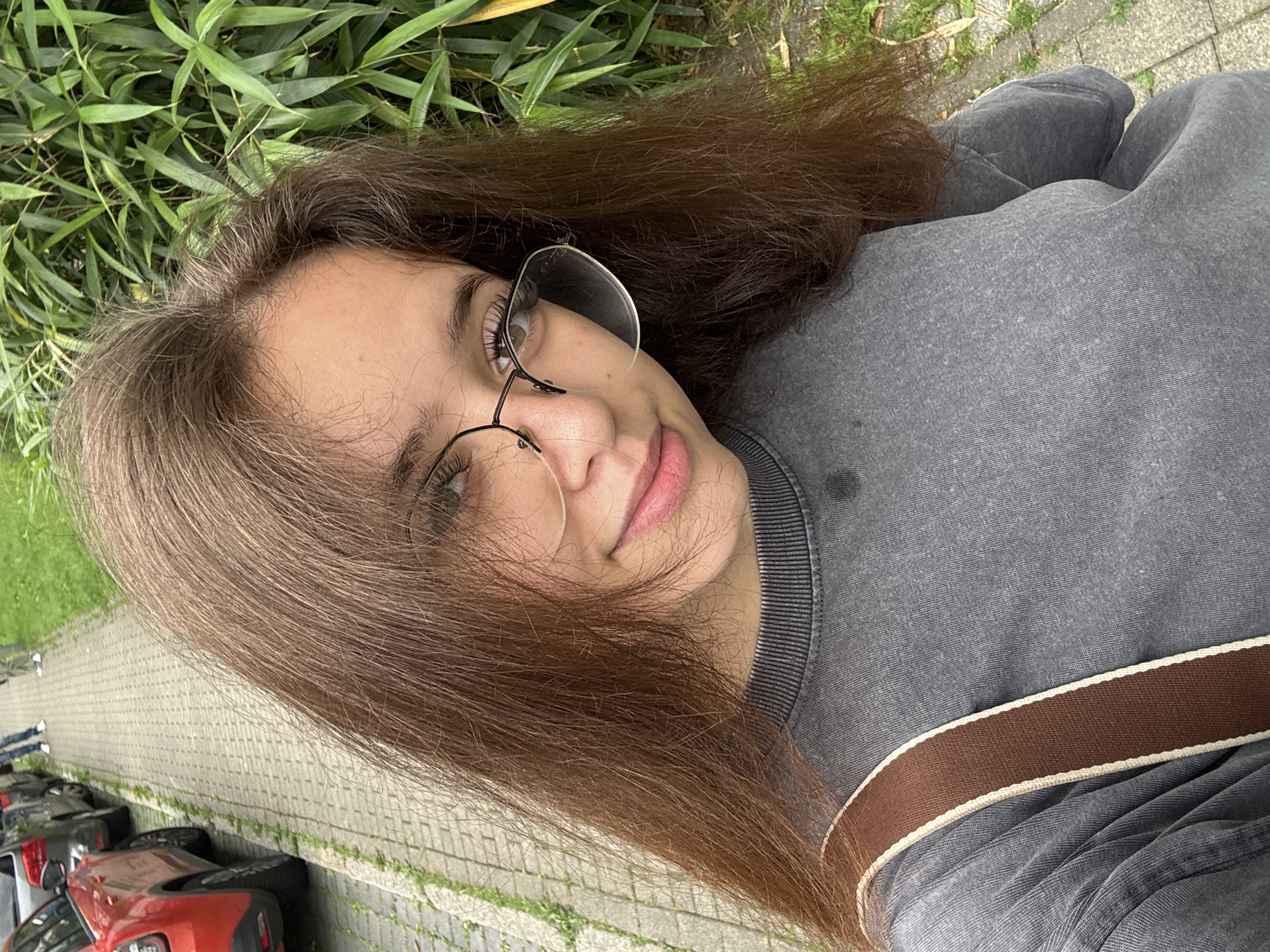 In Göttingen, Germany, a young woman with long, brown hair and glasses is captured in a selfie. She is wearing a grey sweatshirt with a brown strap across her shoulder. The background features a sidewalk, parked cars, and lush green foliage. The scene suggests an outdoor urban setting.