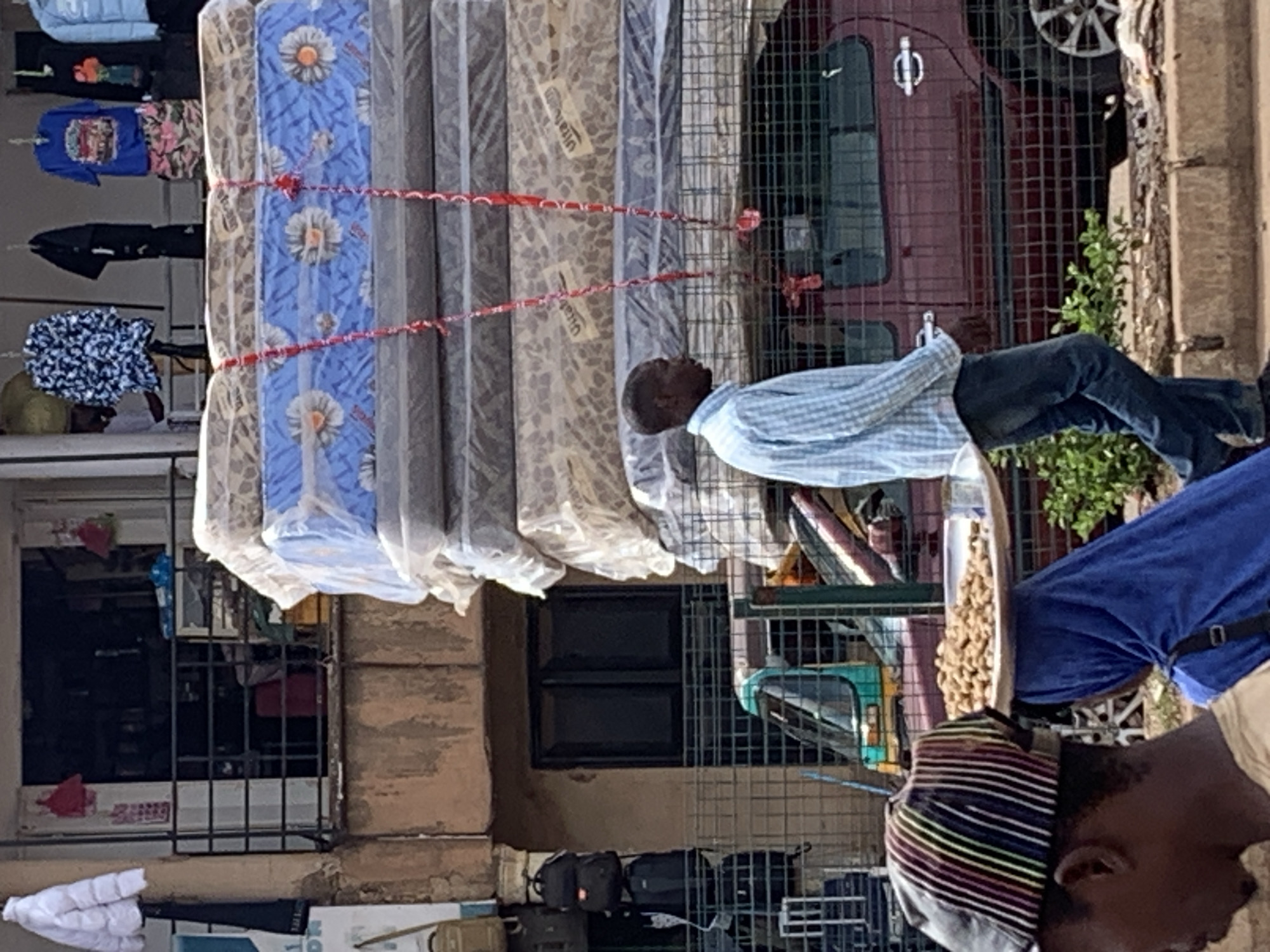In Bukuru, Nigeria, a man in a blue checkered shirt and jeans walks past a stack of mattresses on a flatbed truck. The mattresses are wrapped in plastic and tied with red string. In the foreground, a person wearing a striped cap is partially visible, and behind them, a person in a blue headscarf carries a tray of what appear to be nuts on their head. A green and yellow three-wheeled vehicle is visible behind a metal fence. The background shows a building with a shop entrance and hanging clothes.