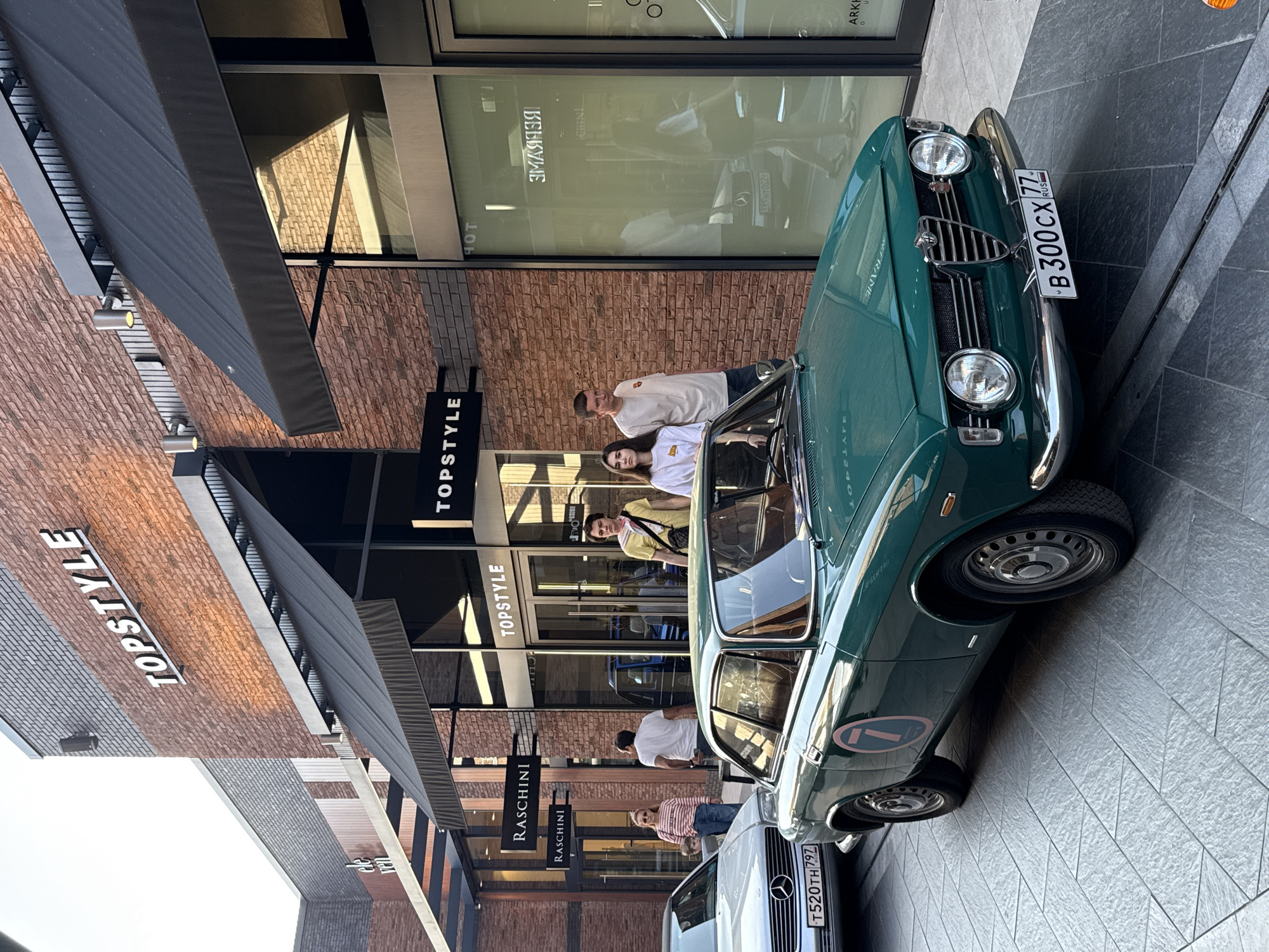 Here is a detailed description of the image:
The image shows a green vintage car parked on a paved area in front of a building with storefronts. The building appears to be a retail space, featuring signs for businesses like "TOPSTYLE" and "RASCHINI." The building has a modern design with brick and large glass windows. Several people are standing near the entrance of the shops, possibly customers or passersby. There are black awnings above the storefronts. A second car, a white Mercedes Benz, is partially visible behind the green car. Both cars have license plates: the green car's plate reads "B 300 CX 77" and the Mercedes's plate reads "T520TH 797." The area is well-lit, suggesting it is daytime. The location is in the city of Unknown, Unknown, based on the Russian license plates on the vehicle.