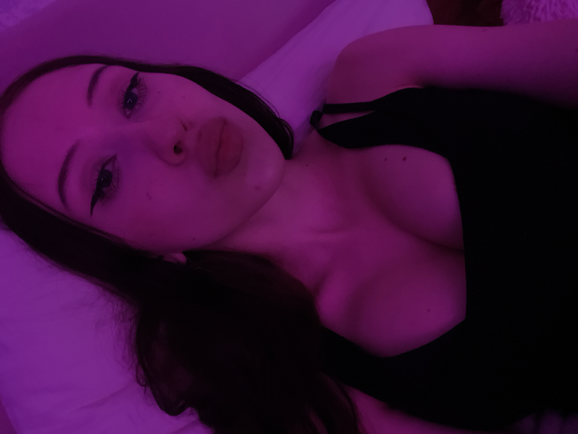 The image shows a close-up, high-angle selfie of a young woman with long, dark hair. She is wearing a black top with a square neckline. Her face is angled towards the camera, and she has dramatic winged eyeliner. Her lips are pursed in a "duckface" pose. The scene is illuminated with purple lighting, giving the image a distinct, artificial glow. The background appears to be a bed with white pillows. The specific location in Votkinsk, Russia, cannot be determined from the visual information provided in the image.