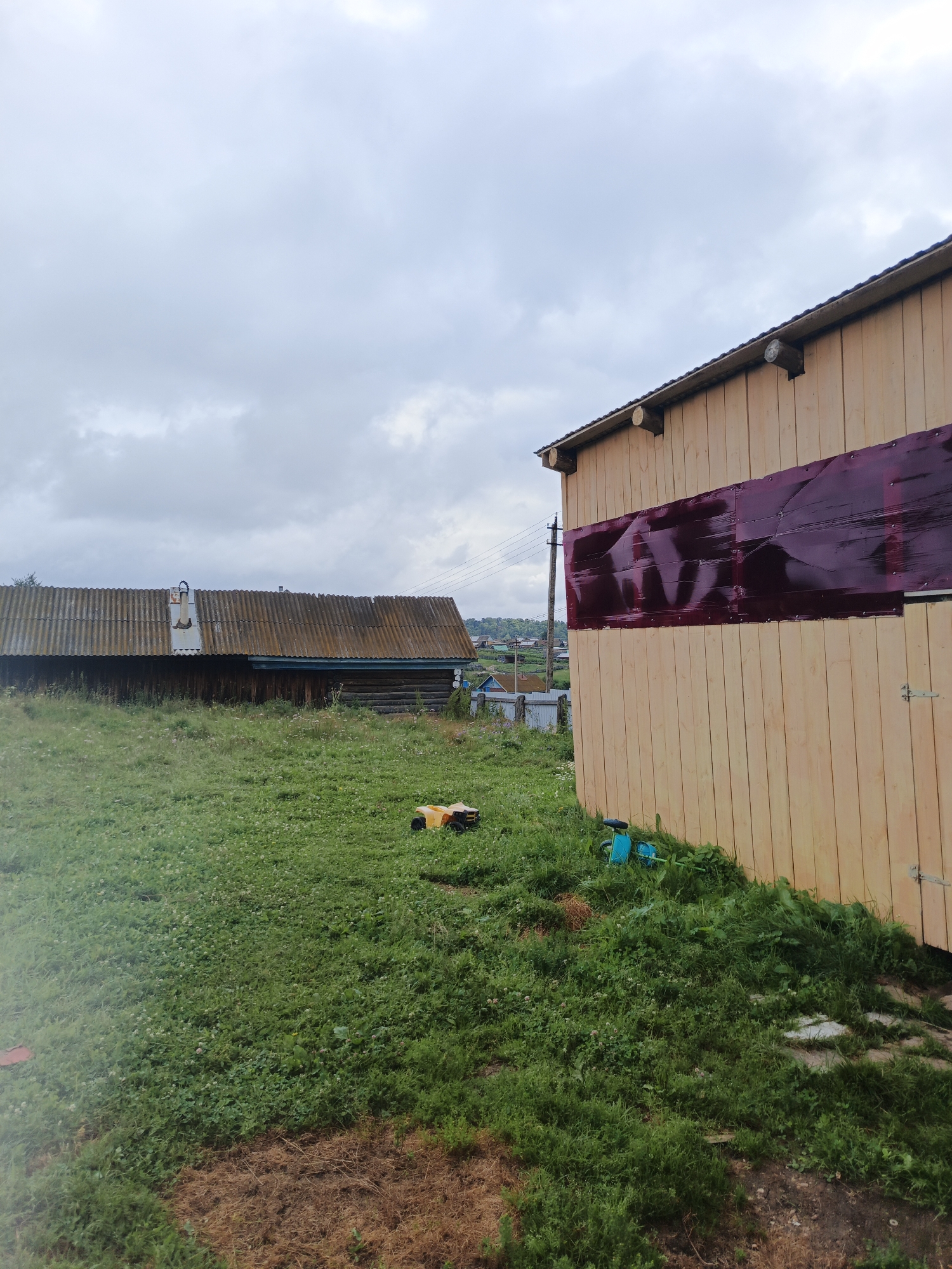 This image depicts an overcast day in Islambaevo, Russia, with a focus on two rural structures and surrounding grassy terrain. In the foreground and right side of the frame is a wooden building under construction, its light-colored vertical planks forming a prominent wall. A deep red, possibly plastic, sheeting is affixed horizontally across the upper section of this building's wall. To the left, a second, older building is visible, characterized by a corrugated metal roof that appears weathered and tinged with moss. This older structure has dark wooden walls.
The ground is covered in lush green grass, with patches of dry, brown earth and some small, scattered white flowers. A yellow toy ATV is parked on the grass in the middle ground, and a blue child's toy, possibly a watering can, lies on the ground near the base of the newer building. In the distance, a cluster of smaller, more traditional rural houses can be seen, suggesting a village or hamlet setting. Electrical poles and wires are also visible in the background, indicating the presence of infrastructure. The sky is filled with dense, gray clouds, contributing to the subdued lighting of the scene. There are no people visible in the image.
