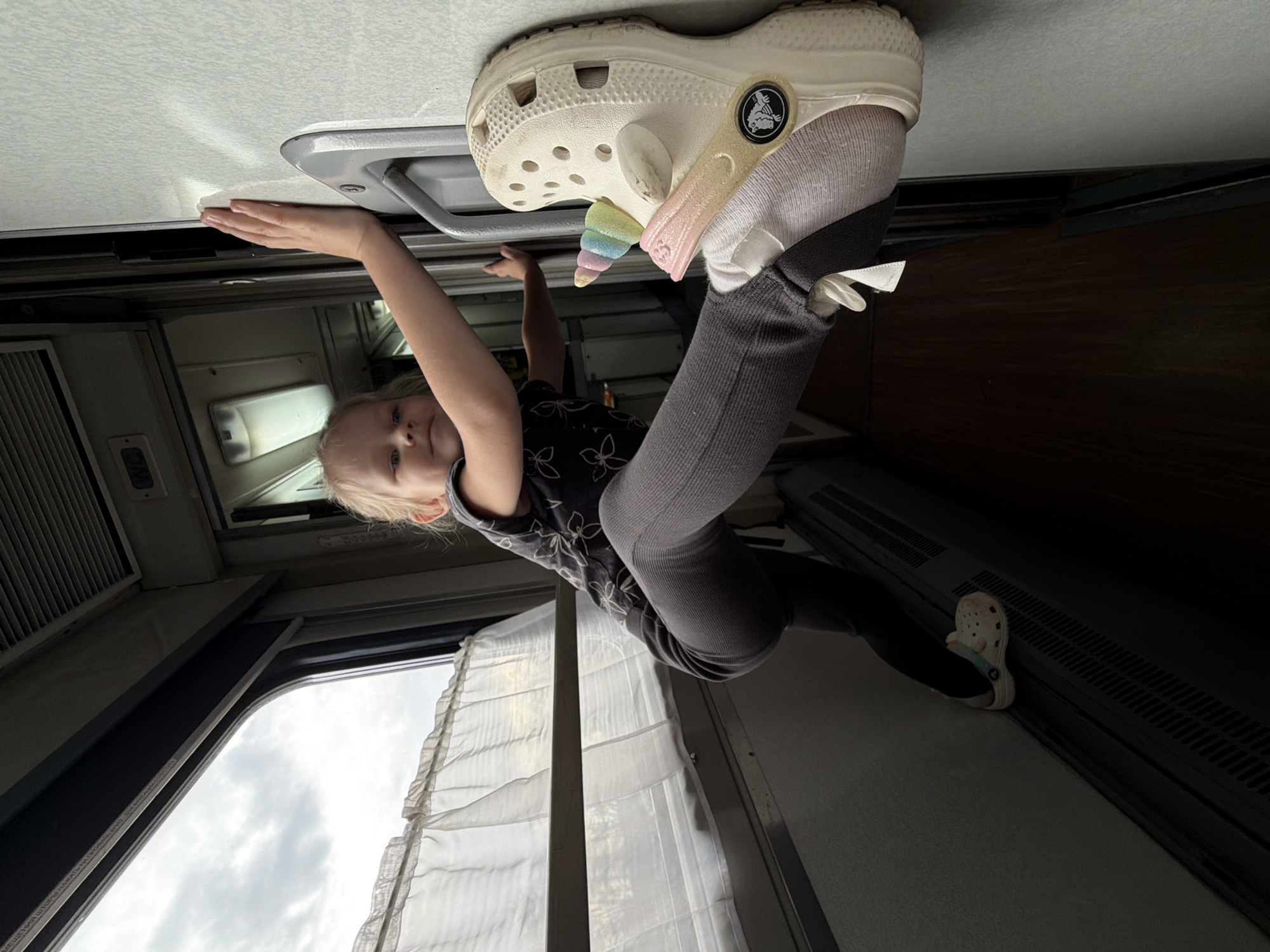 A young child, likely a girl with blonde hair, is depicted hanging upside down in a playful manner inside what appears to be a train carriage or a long-distance bus. Her left hand is gripping an overhead grey handle or fixture on the light-colored ceiling, while her left foot, clad in a white Croc shoe adorned with colorful unicorn and glitter charms, is pressed against the ceiling. Her right foot, also wearing a white Croc with a light blue strap, rests against a dark, slatted panel running along the lower wall of the vehicle.
The child is dressed in a dark patterned short-sleeved top and dark leggings, and looks directly at the camera with a neutral expression. The interior features light-colored walls, overhead lighting, and windows with white, ruffled sheer curtains. Through the windows, an overcast or cloudy sky is visible, indicating daytime. The Crocs brand logo, including the word "CROCS" and the crocodile emblem, is clearly visible on the shoes. The scene captures a lighthearted moment within a transit setting in Urusovo, Russia.