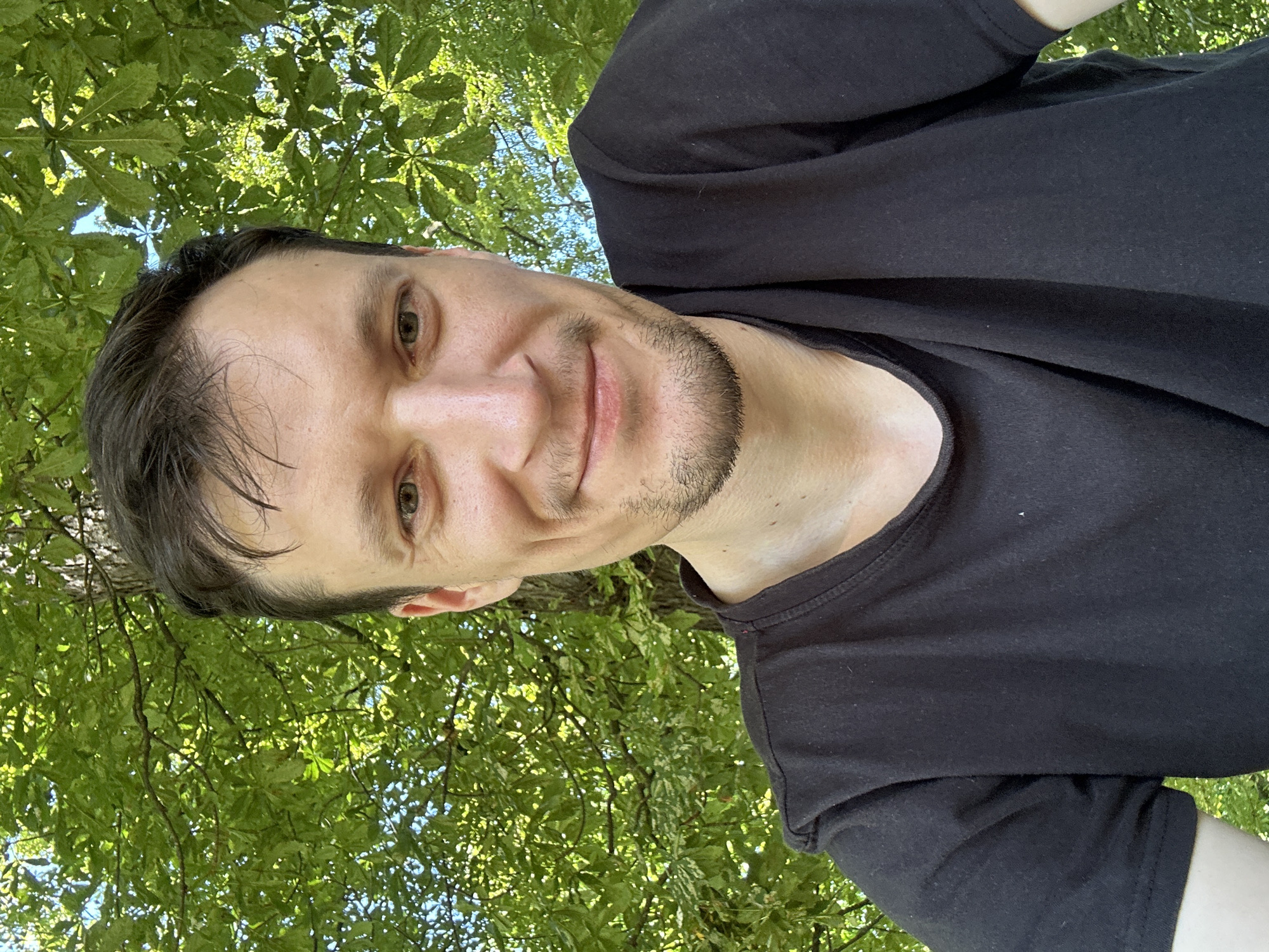 A close-up, outdoor selfie shows a man in his late 20s or early 30s with short, dark hair and a light beard, smiling. He is wearing a black t-shirt. Behind him, lush green leaves and branches of a tree are visible, with dappled sunlight filtering through. The blue sky can be seen in the background. There is no indication of the specific location within Makiv, Ukraine.