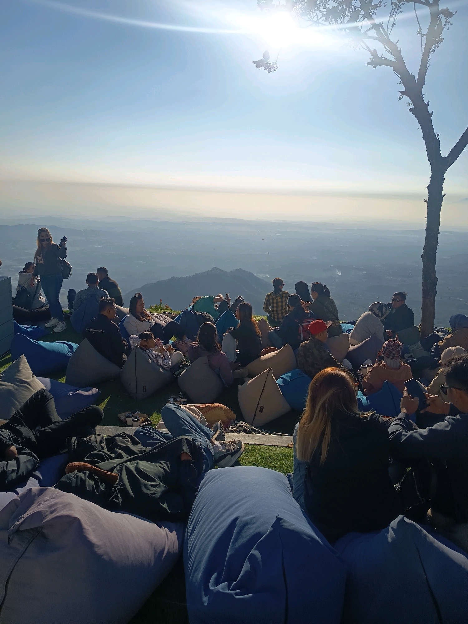 The image depicts a group of people gathered on a grassy hillside overlooking a hazy, mountainous landscape. The scene is bathed in bright sunlight, with lens flare visible in the upper portion of the frame. Many individuals are seated on large, colorful beanbag chairs. Some are interacting, while others appear to be observing the view or using their phones. A lone tree with sparse foliage stands on the right side of the frame, silhouetted against the sky. The location is identified as Keditan, Indonesia.