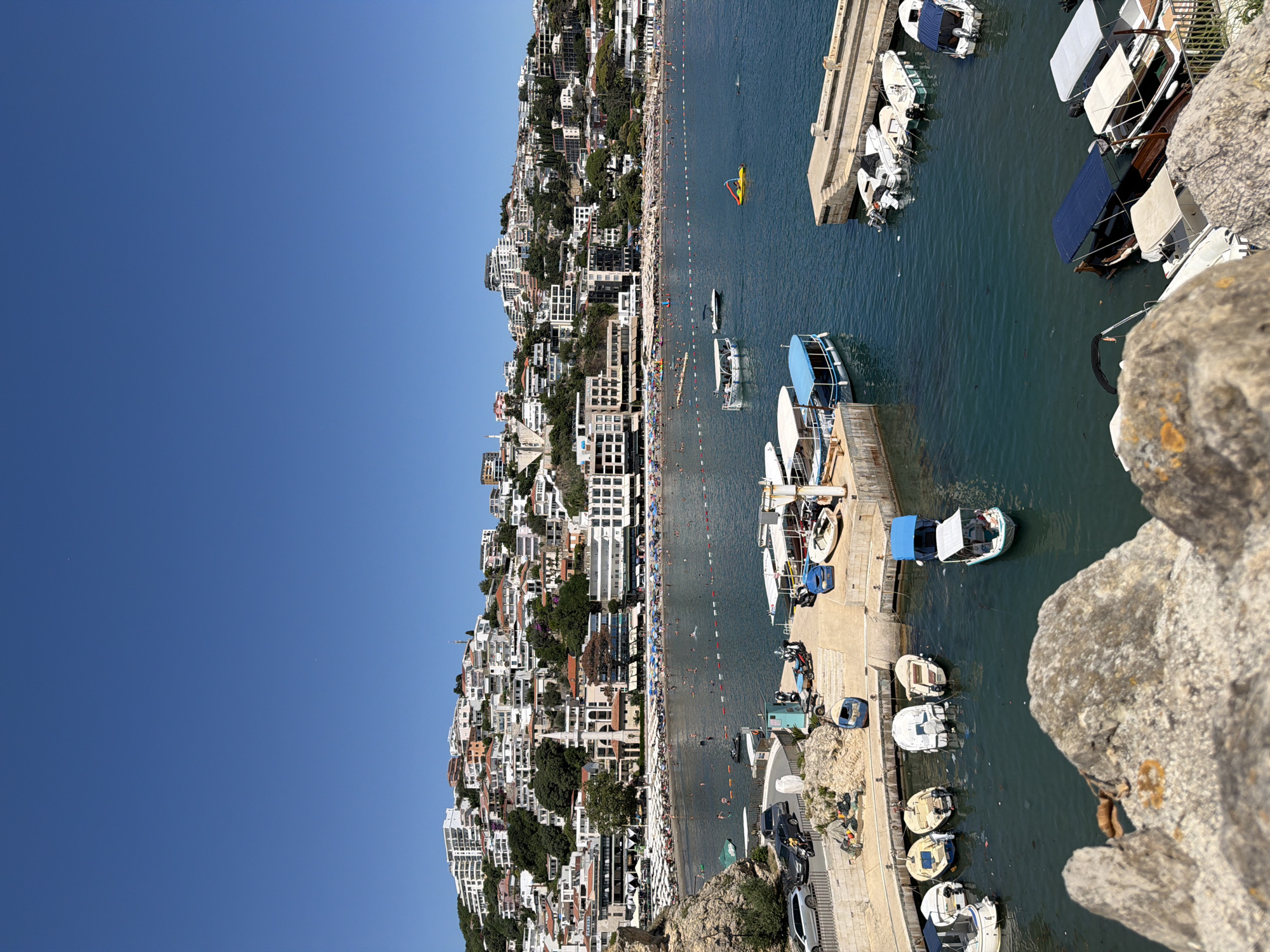 This image captures a panoramic view of a bustling coastal city, identified as Ulcinj - Ulqin, Montenegro. The scene unfolds with a clear, bright blue sky dominating the upper portion of the frame. Below, a dense cluster of white and red-roofed buildings cascades down a verdant hillside, forming the urban landscape. The buildings are characterized by their modern architecture, with balconies and numerous windows visible.
In the foreground, the foreground is occupied by rough, textured grey rocks, partially obscuring the view of the water below. The water itself is a deep, vibrant blue, dotted with numerous small boats. Some boats are moored together at a concrete dock, while others are scattered across the bay. Several of these are small motorboats, some with canopies, suggesting recreational or transport purposes.
Further out in the bay, a long, sandy beach is visible, lined with numerous beach umbrellas and loungers, indicating a significant number of beachgoers. People are also seen swimming and enjoying the water. A few larger vessels are present further out to sea. The coastline is a mix of rocky outcrops and the developed beach area.
The image is taken from an elevated viewpoint, providing a sweeping perspective of the city, the beach, and the harbor. The lighting suggests it is a sunny day, with strong shadows cast by the buildings and boats. The overall impression is one of a lively tourist destination during peak season.