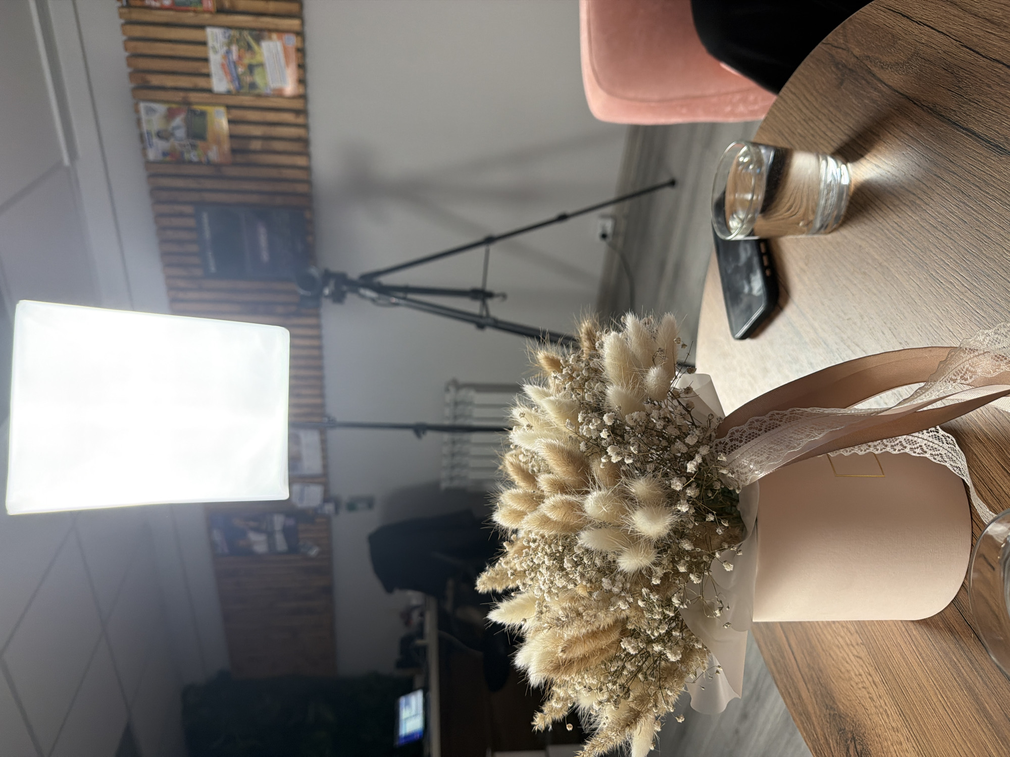 The image shows a bouquet of dried flowers in a pink round box with lace and ribbon accents, placed on a wood-grain table. A glass of water and a black smartphone are also on the table, and a pink chair is partially visible in the background. Behind the table are lighting equipment, including a bright rectangular panel and a tripod. A wall with wooden slat decor, featuring magazine clippings or posters, is also in the background. The location is Chaikovsky, Russia.