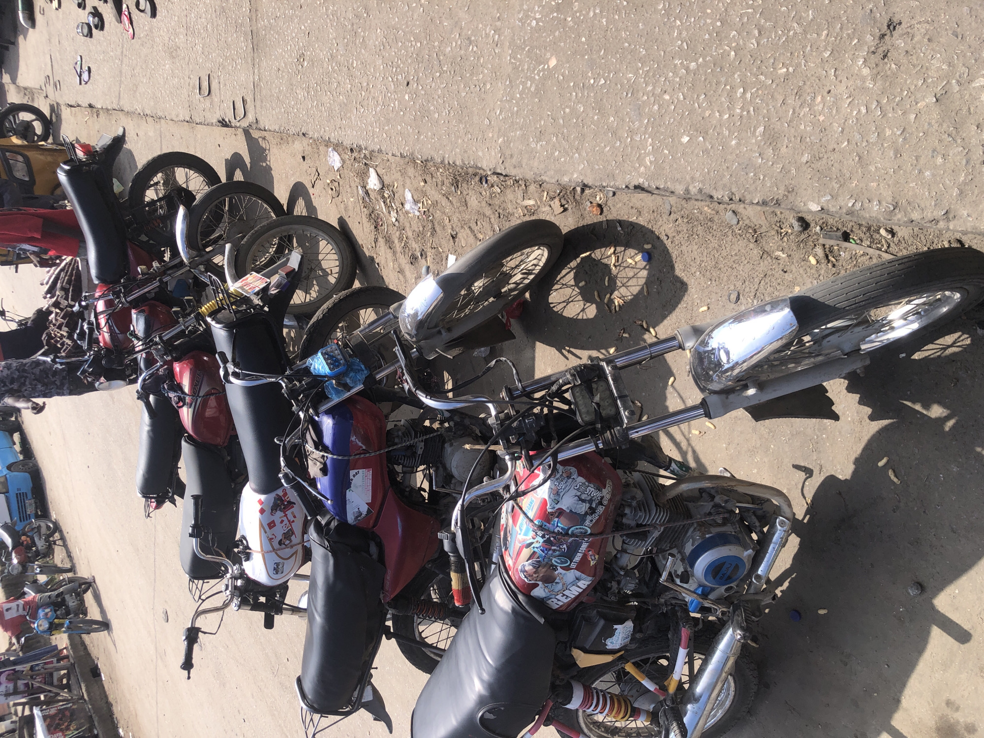 The image depicts a row of motorcycles parked on a dirt shoulder next to a paved sidewalk. The scene is likely in Ado, Nigeria, given the context. Several motorcycles are visible, with the closest one being a red and white model adorned with various decals and stickers. The motorcycles are packed closely together, suggesting they are either for sale or being used for local transport. In the background, there are a few individuals walking and some vehicles, including a yellow auto-rickshaw and a blue truck, indicating a busy urban environment. The ground is dusty, and scattered debris, including plastic bags and what appear to be peanut shells, are present. The overall impression is of a bustling street scene, common in Nigerian cities.