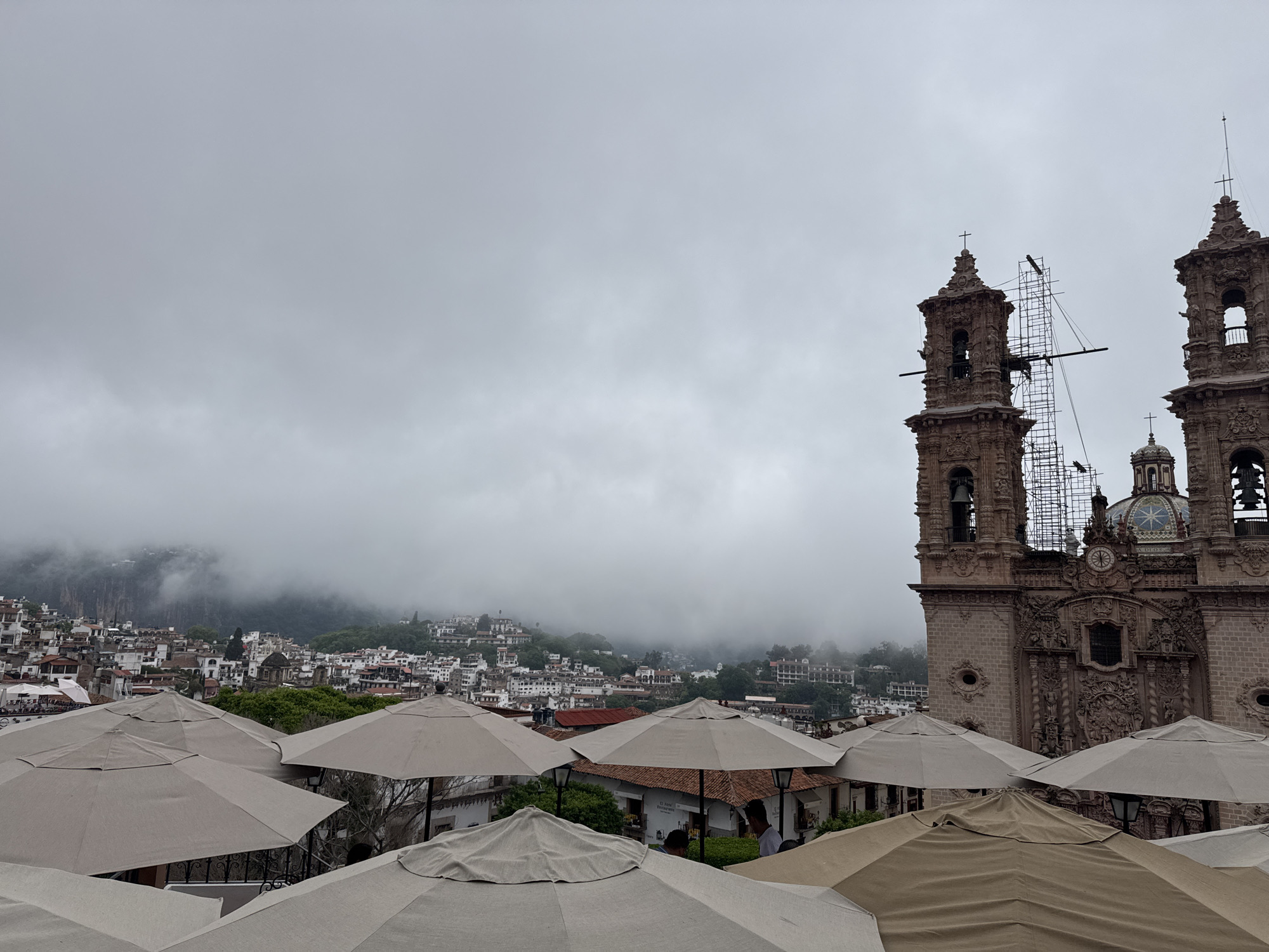 This image captures a view of Taxco, Mexico, under an overcast sky. In the foreground, several large, light-colored umbrellas provide shade, suggesting an outdoor seating area, possibly a restaurant or cafe. Behind the umbrellas, the distinctive architecture of Taxco unfolds. To the right, a large, ornate church with twin bell towers dominates the scene. One tower is partially obscured by scaffolding, indicating ongoing renovation or maintenance. The church is constructed of reddish-brown stone and features intricate carvings and multiple crosses atop its spires. In the middle ground and background, a dense collection of white-and-terracotta buildings cascades down a steep hillside, characteristic of Taxco's hillside setting. The upper parts of the hills are shrouded in mist, adding a dramatic and somewhat ethereal quality to the landscape. The overall impression is a blend of urban and natural scenery, with a prominent religious structure overseeing a town nestled within a mountainous environment.