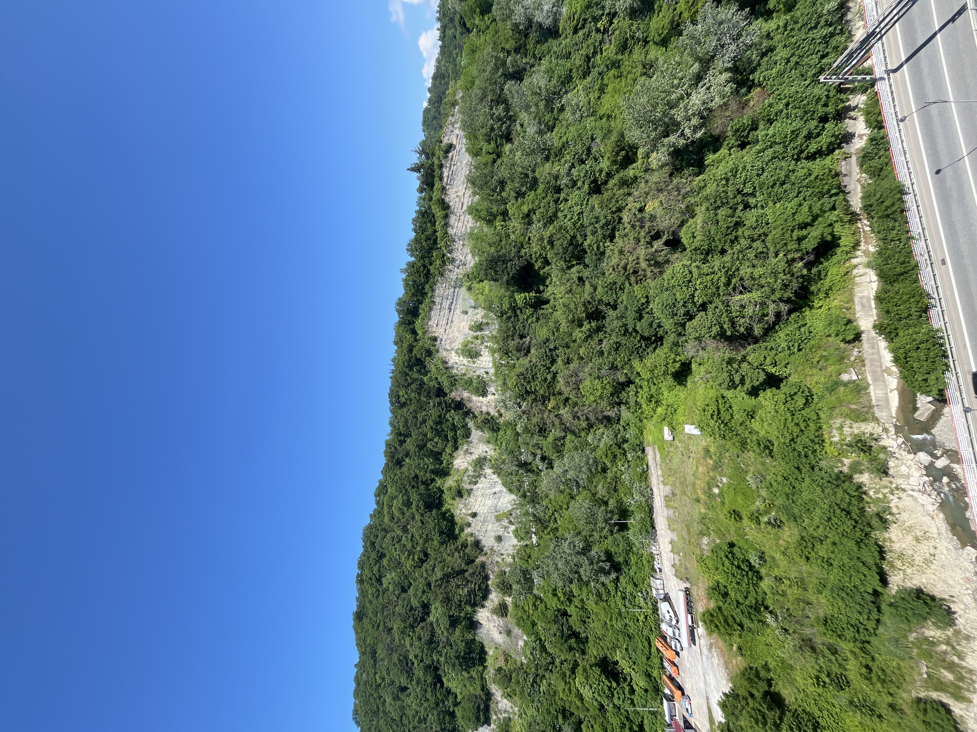 This is a high-angle, wide shot of a mountainous area with a bright blue sky overhead. The mountainside is densely covered in lush green trees, with a visible rocky cliff face emerging from the foliage. At the base of the mountain, there is a flat area with several vehicles parked, including orange and white trucks and trailers. Below this, a body of water, appearing to be a river or stream, flows between rocky banks. A road with a guardrail runs along the lower portion of the image, parallel to the water. The scene suggests a natural, undeveloped area on the outskirts of a city, likely Sochi, Russia, given the mountainous terrain and presence of a highway. No people are visible in the frame.