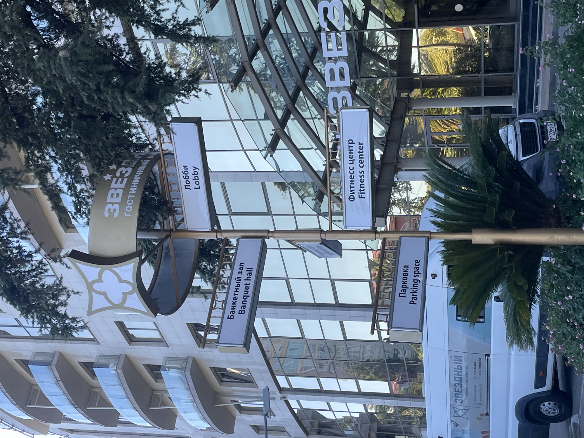 The image shows a signpost outside a hotel in Sochi, Russia, with signs pointing to various amenities. The hotel's facade is a modern building with curved balconies and large glass windows. Trees and foliage surround the entrance. The signs on the post are in both Russian and English, indicating directions to the "Lobby," "Banquet hall," "Fitness center," and "Parking space." A white van with the hotel's logo is parked nearby, and a dark-colored car is visible in the background. The overall scene suggests a well-maintained tourist or accommodation facility.