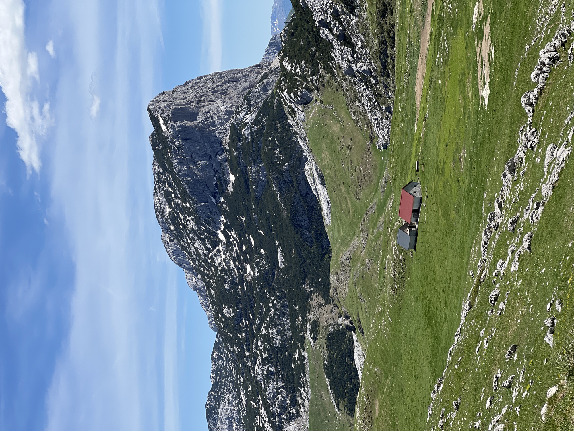 The image shows a landscape in Dubrovsko, Montenegro. The dominant feature is a large, rocky mountain with patches of snow and vegetation clinging to its slopes. To the right of the mountain, a steep, grassy hillside slopes down to a small cluster of buildings with red and grey roofs. The sky above is blue with streaks of clouds. The scene is bathed in daylight.