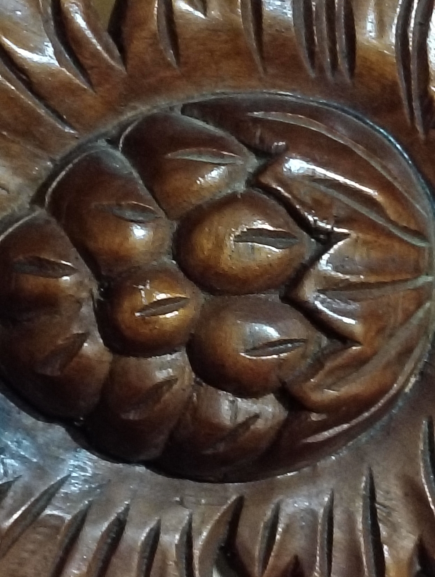 The image shows a close-up view of an intricate wooden carving. The carving appears to be a floral motif, possibly a sunflower, with detailed petals radiating outwards. In the center of the carving, there are several bulbous shapes that resemble seeds or a central core. These shapes are also carved with subtle details that suggest a texture or pattern. The wood itself has a rich, dark brown color and a glossy finish, indicating it is likely polished or varnished. The lighting in the image highlights the texture and depth of the carving, creating shadows and reflections that emphasize the craftsmanship. The image does not contain any people, actions, or interactions, and the environment is not visible. Without further information, it is not possible to determine the specific location of this carving in Payagaboh, Indonesia.