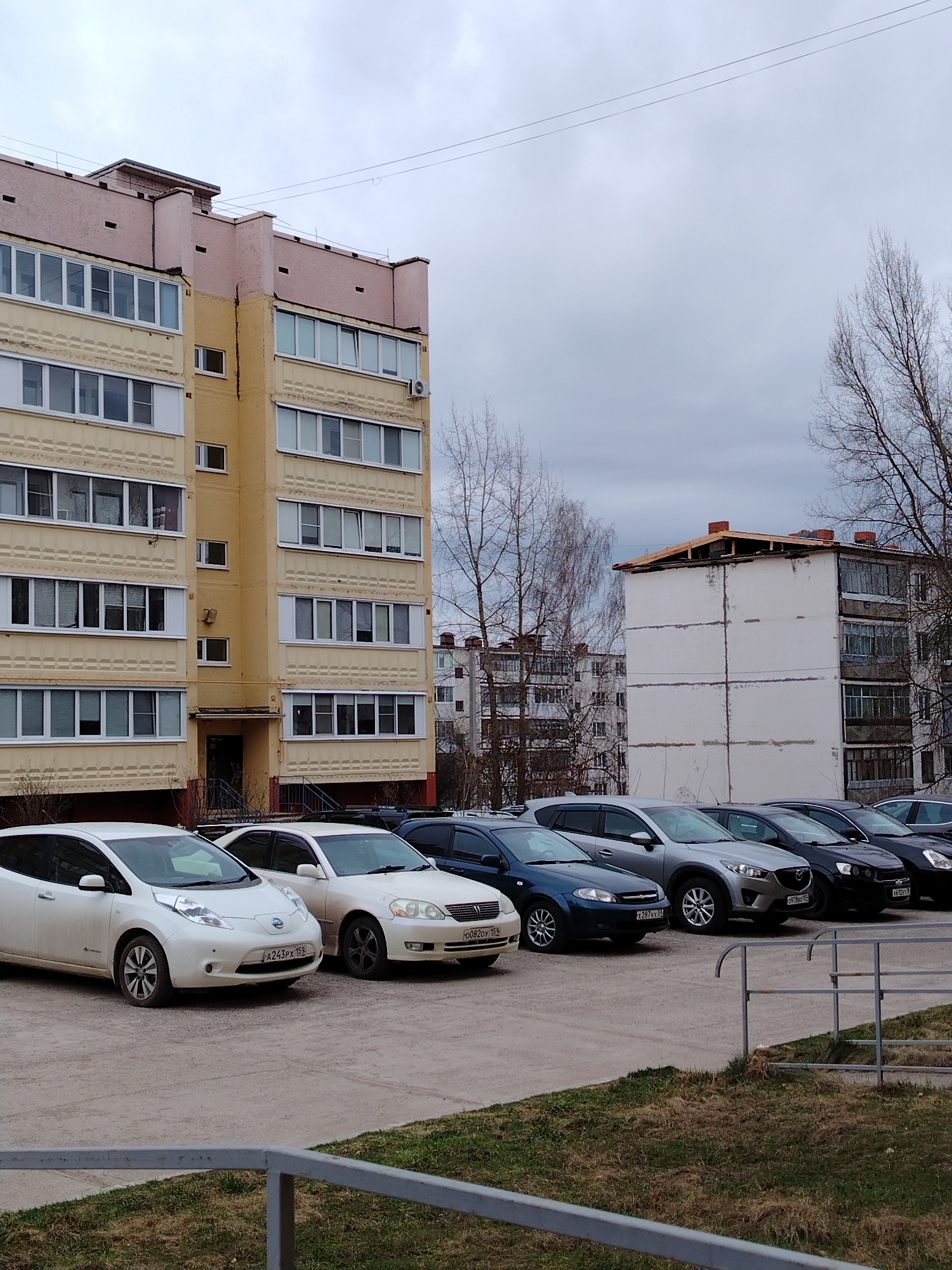 The image shows an outdoor scene in Noviy, Russia. A row of parked cars occupies the foreground of the scene. The cars range in color, with white and dark hues dominating. The license plates on the cars are visible. Behind the parked cars are two residential buildings with multiple floors and windows. The building on the left is painted yellow with pink accents. The building to the right is white. A cloudy sky is overhead. Bare trees are visible in the background. The environment appears to be a parking lot or residential area.