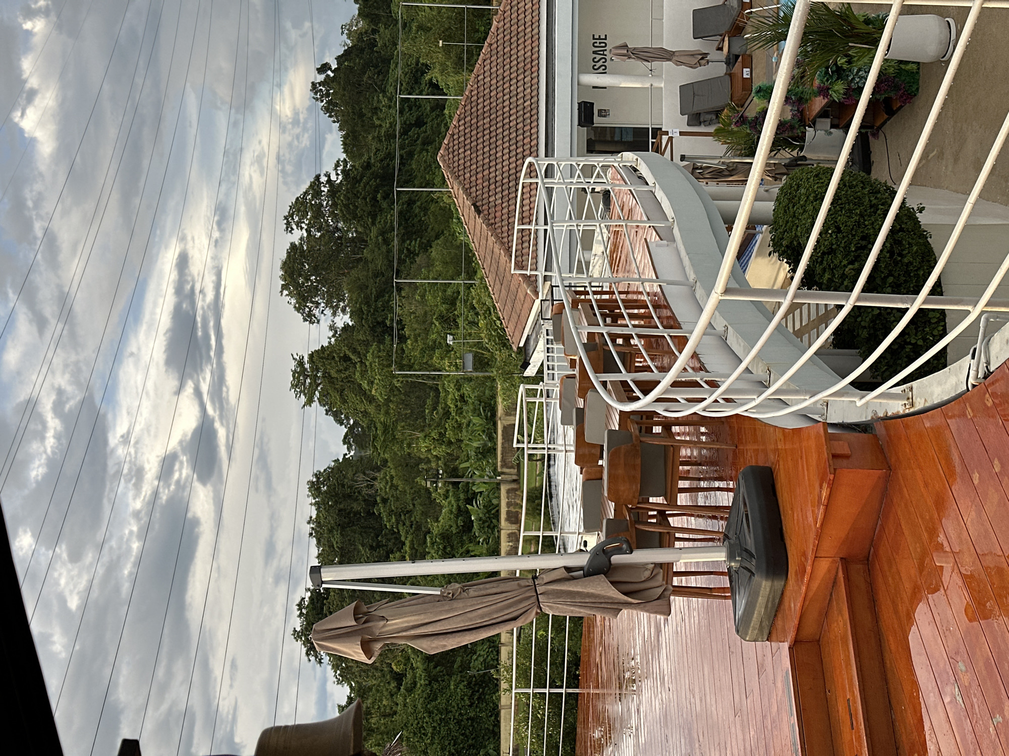 A high-angle exterior shot captures a wet, wooden deck with chairs and tables set up. A closed, tan umbrella stands prominently on the deck. The deck curves around a building with a red tile roof. The building has a sign that reads "MASSAGE." Beyond the building, lush green trees form a backdrop. The sky is overcast with visible power lines stretched across it. In the foreground, railings line the curved pathway leading to the building. The scene suggests an outdoor seating area, possibly at a resort or restaurant, experiencing or having recently experienced rain. The location is the city of Kathu, Thailand.