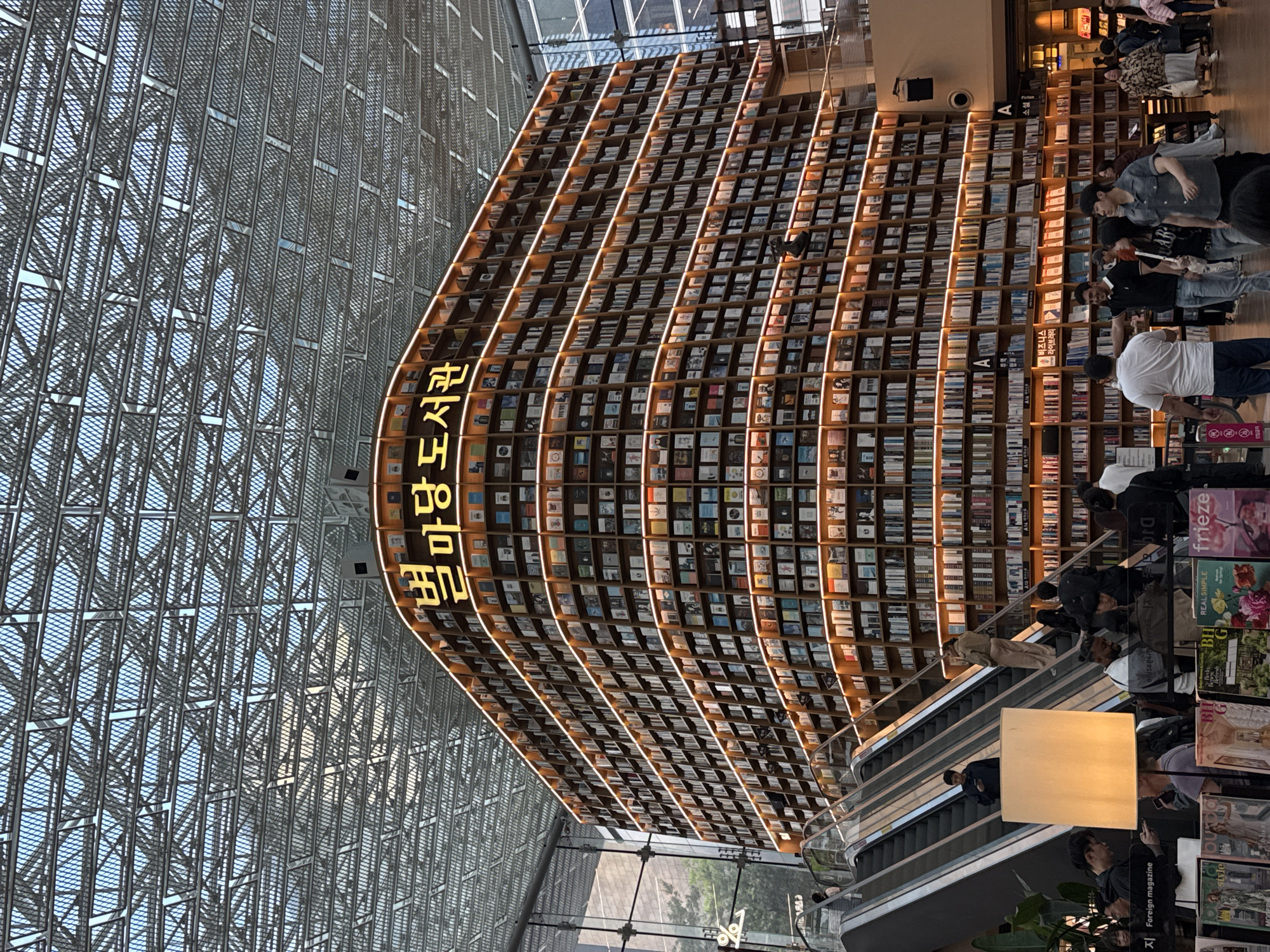 The image shows a tall, curved bookshelf in the Starfield COEX Mall, Seoul, South Korea. The bookshelf, spanning several stories, is filled with books and illuminated by vertical lines of light. A yellow sign reads "별마당 도서관," which translates to "Starfield Library." People are seen browsing the books and standing near an escalator. The backdrop features a large glass and metal grid structure, providing natural light. There are magazine racks on the bottom right, with "Foreign magazine" listed on one.
